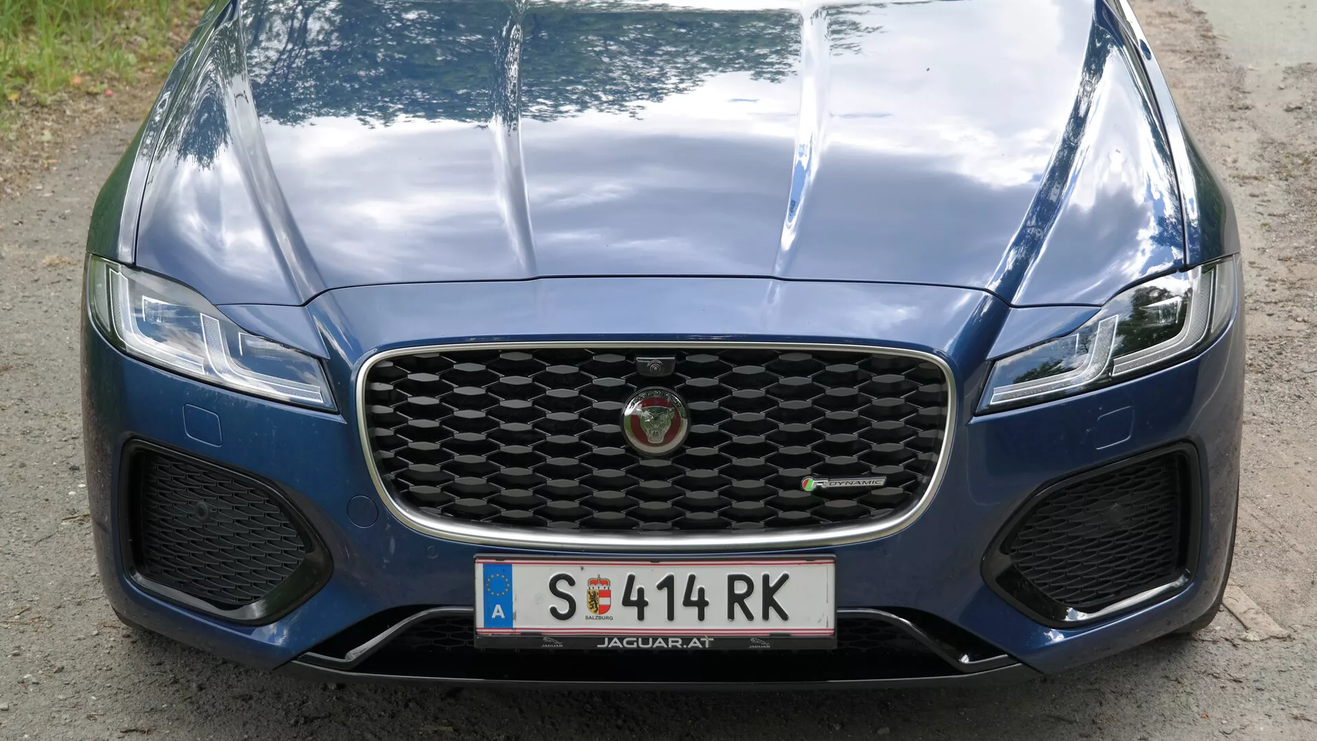 Vista frontale di un'auto di lusso, ripresa da un'angolazione leggermente bassa, evidenziando la griglia, il paraurti e il cofano. La griglia presenta un grande design a nido d'ape, mentre i fari stretti e sottili suggeriscono una tecnologia di illuminazione avanzata. Linee di contorno e una sezione centrale rialzata sul cofano suggeriscono un design aerodinamico. Il paraurti anteriore include grandi prese d'aria, forse per alloggiare i fendinebbia. Un badge distintivo al centro della griglia indica lo stato di lusso del veicolo. La vernice blu metallica è lucida, con riflessi visibili che mostrano l'ambiente circostante. Il primo piano si concentra sul design e sulle caratteristiche di lusso del veicolo.