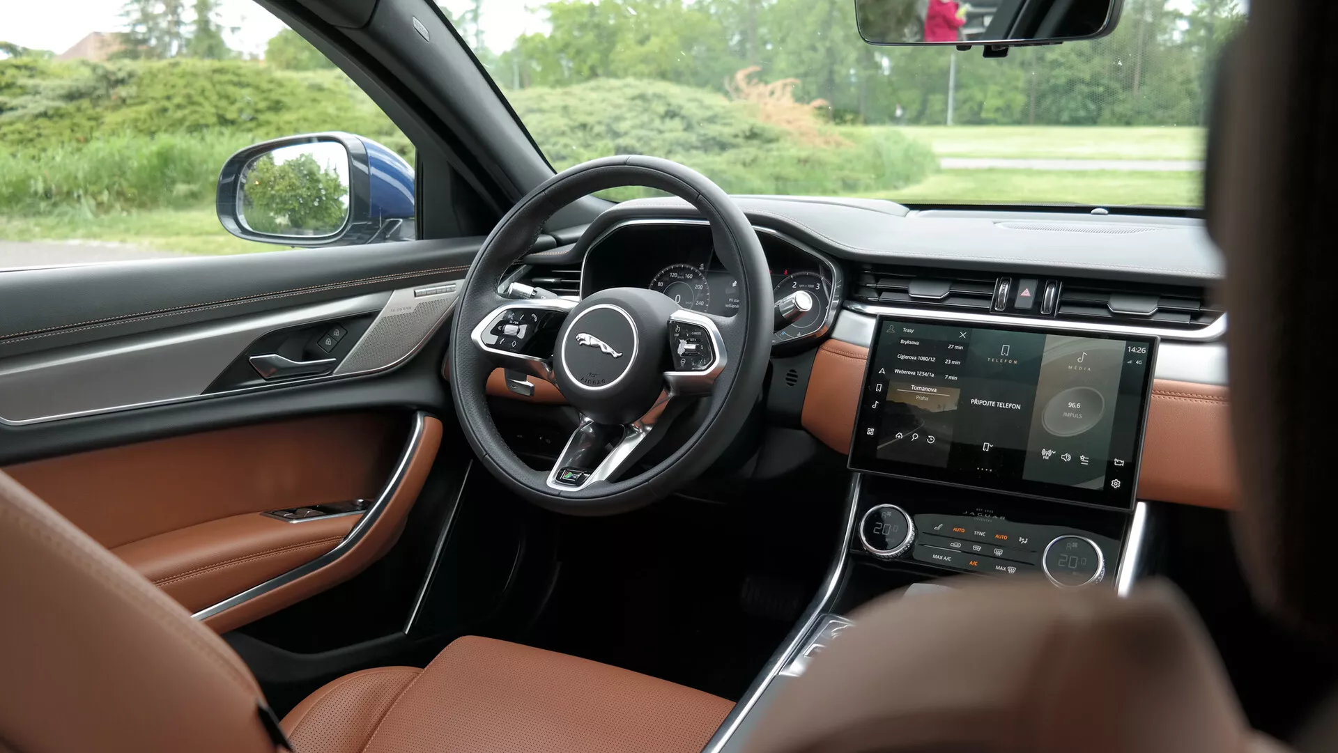 Vista interna di un'auto moderna dalla prospettiva del sedile posteriore, leggermente spostata verso il lato del conducente, che mostra il cruscotto anteriore, il volante, la console centrale e la porta del conducente. Il volante a tre razze ha pulsanti multifunzione su entrambi i lati. Dietro il volante è visibile un quadro strumenti digitale che indica display configurabili per le informazioni del conducente. La console centrale ospita uno schermo tattile per la navigazione, i media e le impostazioni del veicolo, insieme a pulsanti fisici che probabilmente controllano il clima e altre impostazioni. L'interno contrasta il cruscotto scuro con i sedili in pelle marrone chiaro di lusso. La porta del conducente include controlli per i finestrini, gli specchietti laterali e le regolazioni del sedile, indicando un pacchetto tecnologico avanzato. Il veicolo è fermo, circondato da alberi, come si vede attraverso il finestrino laterale, enfatizzando la chiarezza dei dettagli interni.