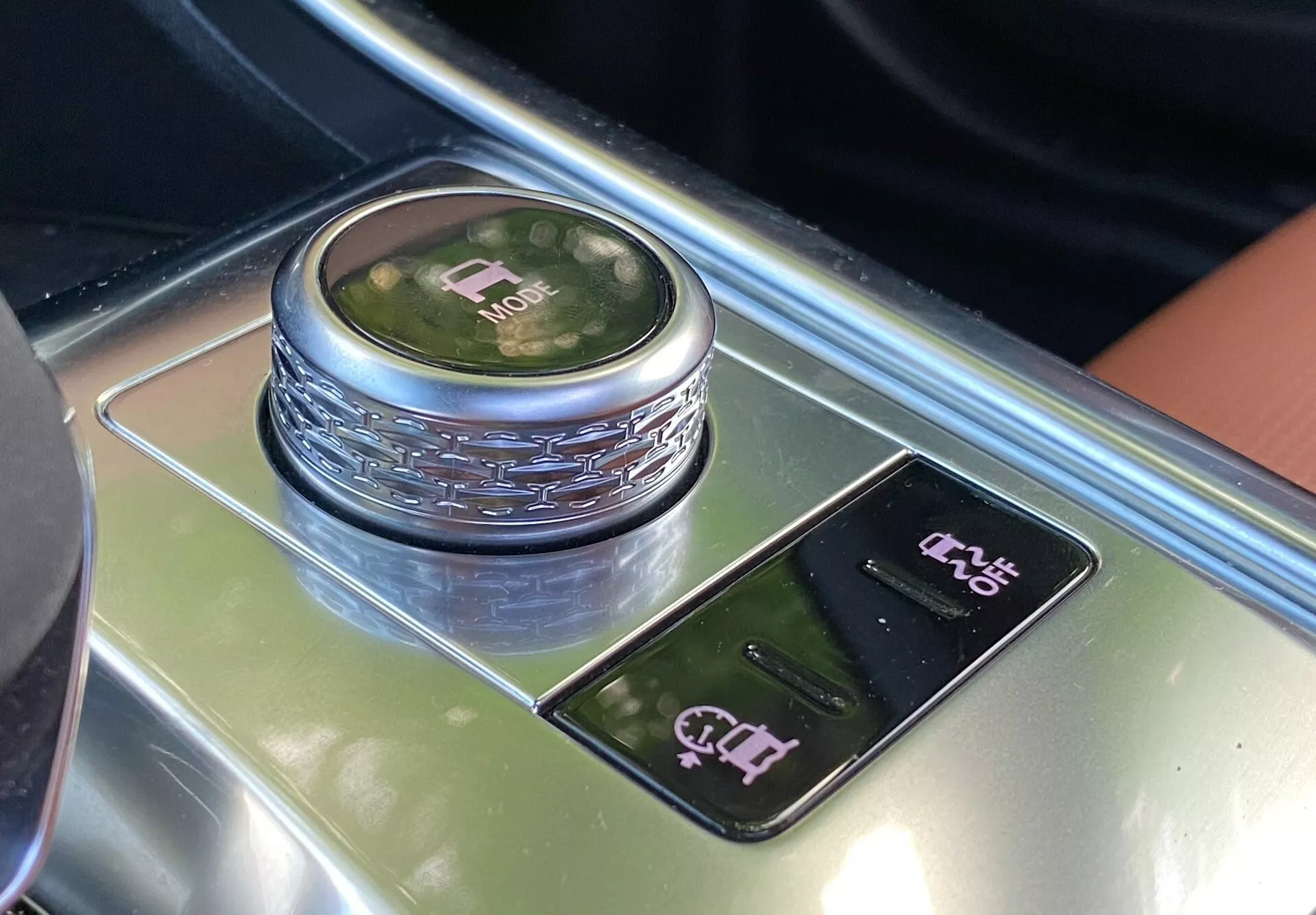 Questa immagine cattura una sezione dell'interno della Jaguar XF 2021, focalizzata sull'area della leva del cambio da una prospettiva dall'alto. La caratteristica principale è una manopola di cambio circolare con una finitura metallica riflettente e un display illuminato che indica la modalità 'P'. Intorno alla manopola c'è un dettaglio decorativo e sotto di essa un pannello lucido con comandi sensibili al tocco, inclusi un controllo elettrico del freno di stazionamento e un altro possibile controllo del sistema di assistenza al parcheggio. I materiali lussuosi, come superfici nere lucide e forse legno o metallo spazzolato, esemplificano l'estetica di design premium di questo interno.