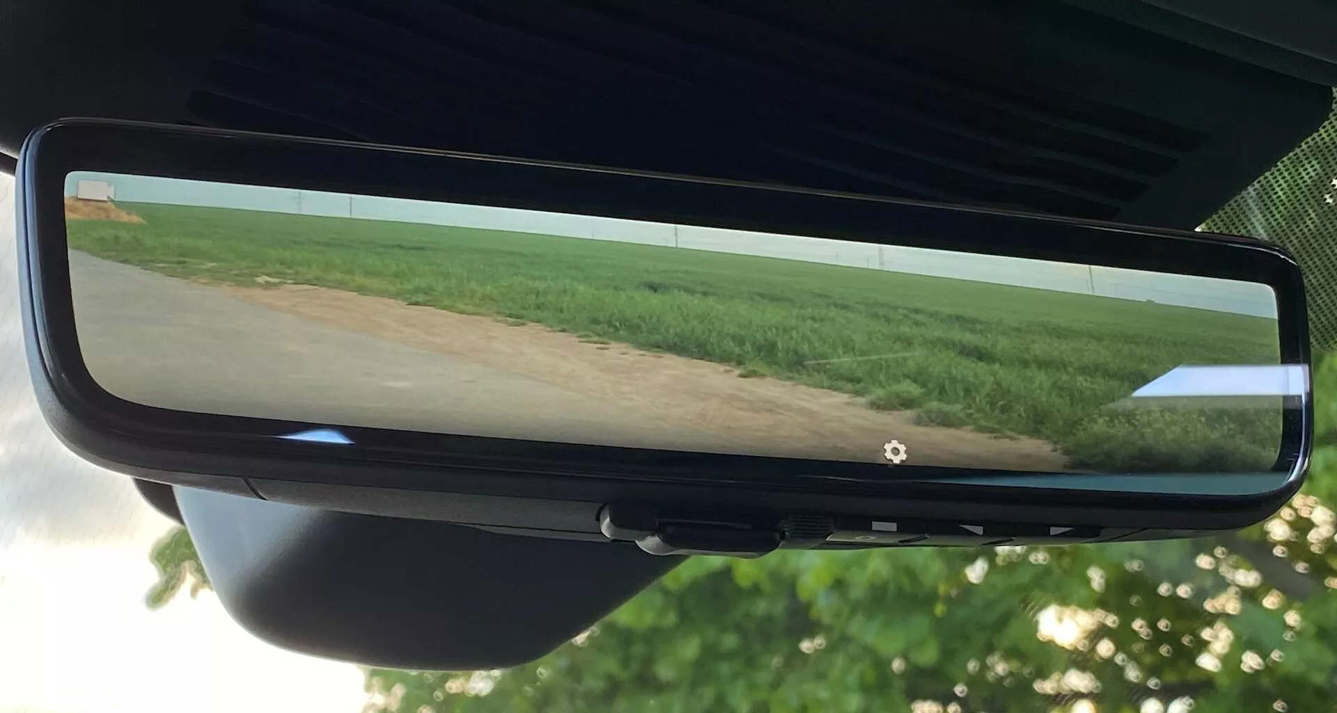 Specchio retrovisore della Jaguar XF 2021 che riflette una vista grandangolare di un campo con vegetazione e cielo limpido, centrato nella foto. Lo specchio è fissato al supporto del parabrezza ed è circondato da una cornice nera, con sezioni del rivestimento interno e del retrovisore visibili.