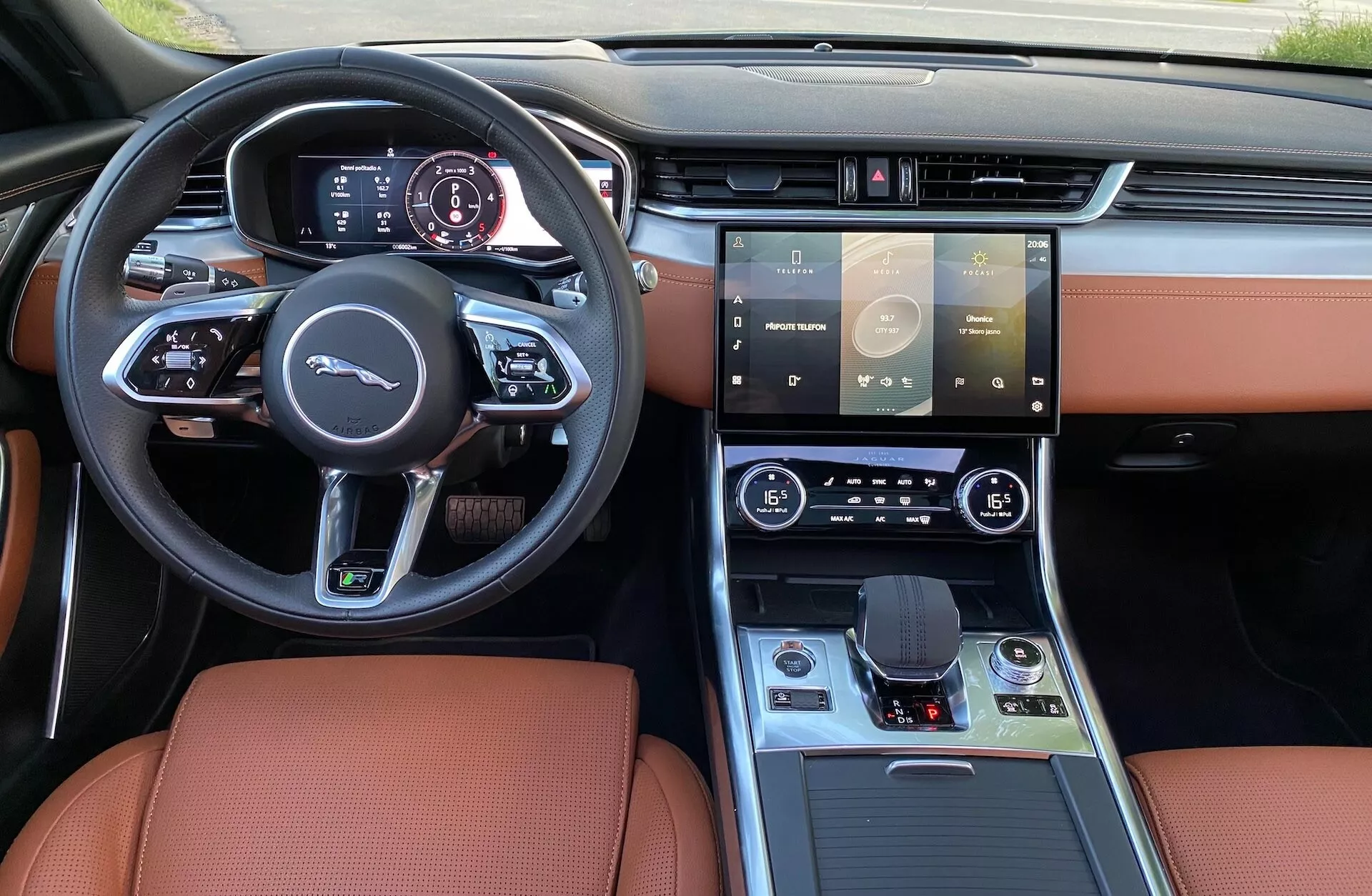 Vista interna della Jaguar XF 2021 dal posto di guida che mostra il volante sportivo con fondo piatto con pulsanti multifunzione, pannello strumenti digitale, ampio sistema di infotainment con schermo touchscreen, comandi per il clima a doppia zona, pulsante di avviamento senza chiave, selettore del cambio elettronico, console centrale moderna con finiture in legno e metallo, e lussuosi rivestimenti in pelle color caramello.