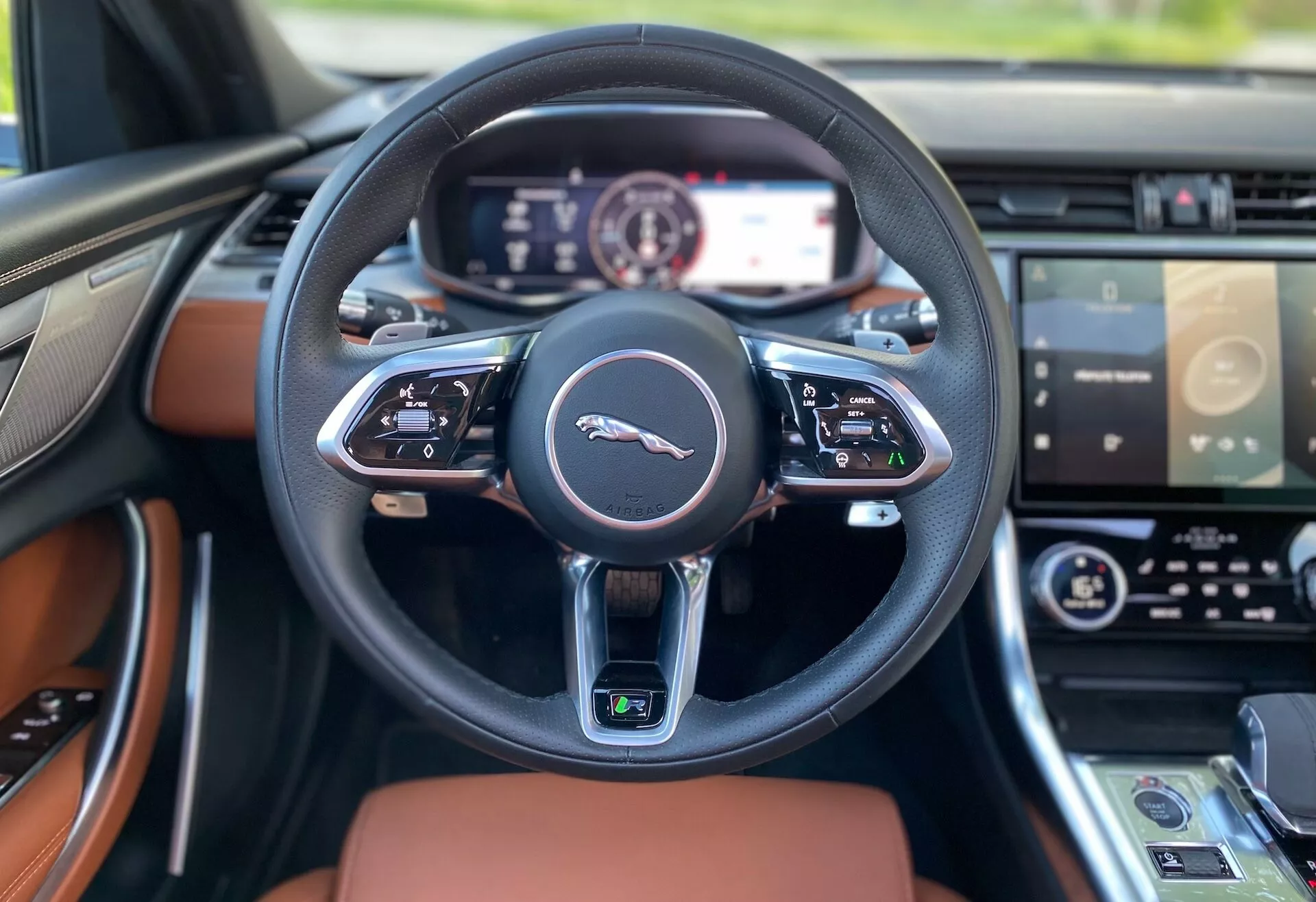 Vista interna della Jaguar XF 2021 dal sedile del conducente, che mostra un volante multifunzione rivestito in pelle nera con un emblema argento e pulsanti di controllo avanzati. Il cruscotto digitale completo dietro il volante presenta un display ad alta risoluzione. Il cruscotto, con inserti in legno di lusso, si estende verso i lati. In secondo piano, è visibile il bordo superiore del sistema di infotainment touchscreen e del pannello di controllo del clima, indicando le caratteristiche avanzate di tecnologia e lusso del veicolo.