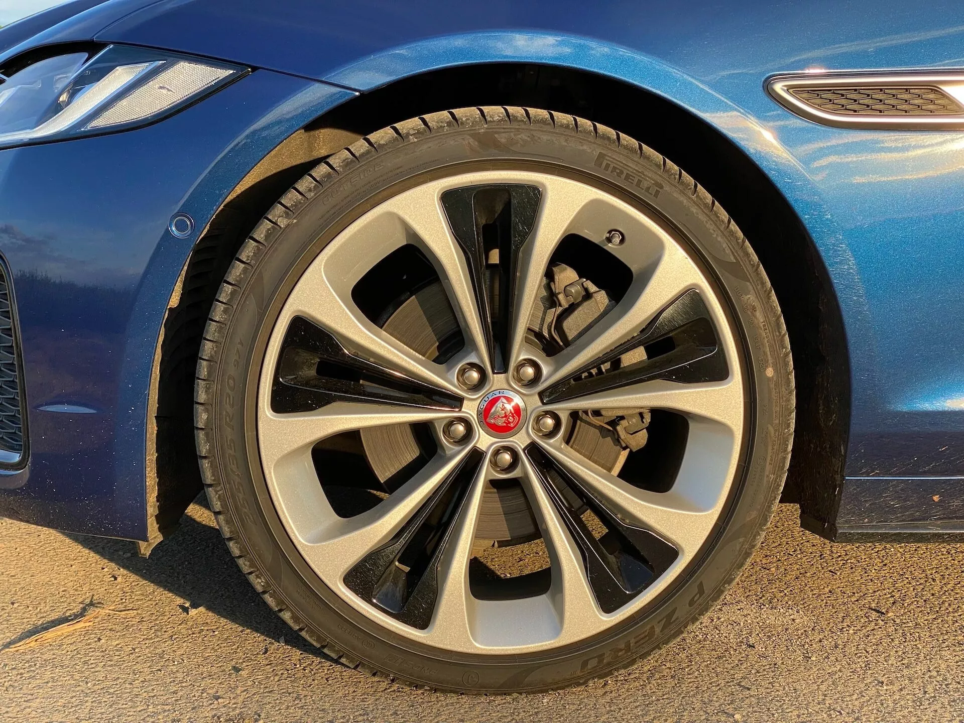 Vista ravvicinata della ruota anteriore lato passeggero di una Jaguar XF 2021 da un angolo basso. La ruota in lega multirazze presenta un design dinamico con dieci coppie di raggi sottili e spessi. Il tappo centrale mostra un emblema rosso della marca. Il pneumatico da strada presenta un visibile schema del battistrada, e dietro i raggi si possono vedere la pinza dei freni e il disco ventilato. È visibile parte della carrozzeria blu con una finitura lucida e il parafango curvo. L'immagine mette in evidenza le caratteristiche di lusso e stile dell'auto, adatta per discussioni su parti di auto, dettagli di design e identificazione del marchio.