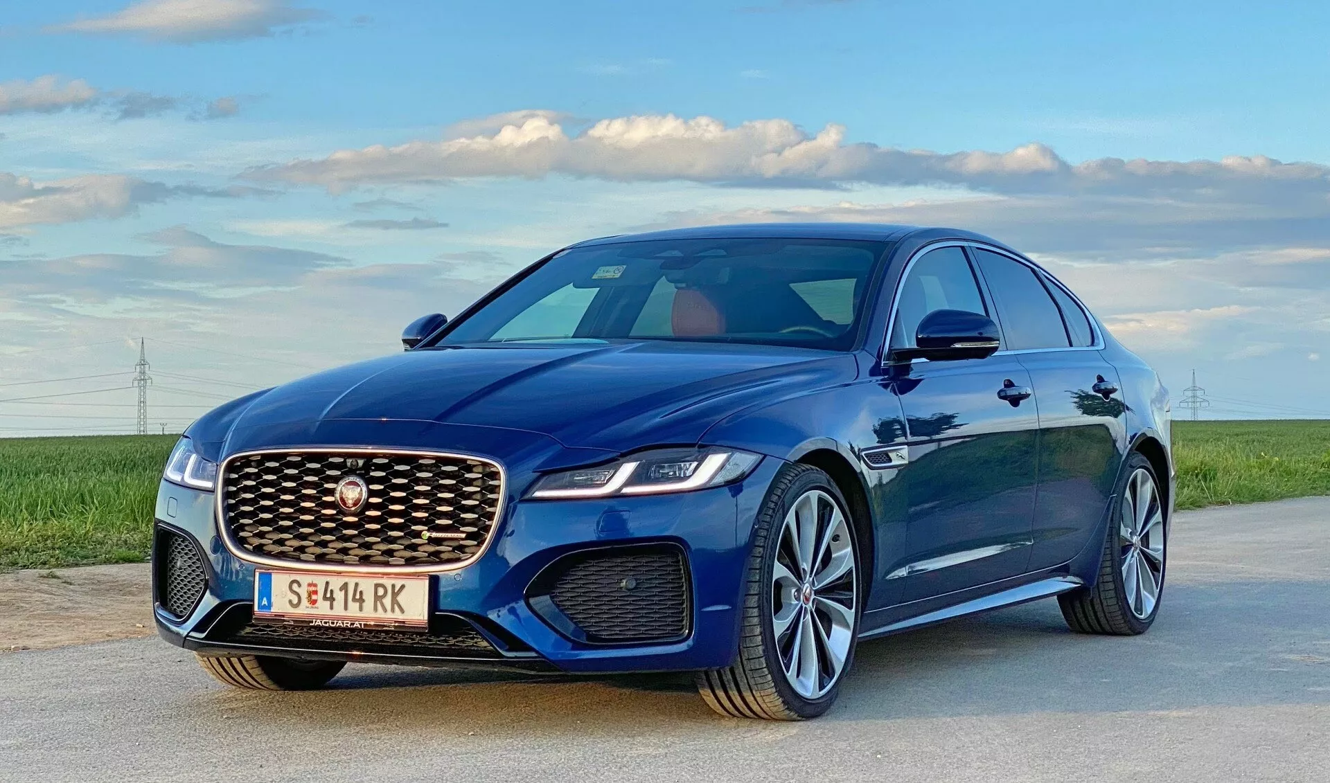 L'immagine mostra una berlina di lusso moderna, una Jaguar XF 2021, parcheggiata su una strada aperta sotto un cielo limpido con campi e linee elettriche distanti all'orizzonte. Catturata da un'angolazione bassa e una vista tridimensionale frontale, sottolinea il lato del conducente. Evidenzia la griglia anteriore prominente con un motivo a maglia e un emblema centrale, i fari a LED eleganti, le linee scolpite del cofano, gli specchietti laterali aerodinamici e i cerchi in lega a razze multiple con vernice metallica. È possibile dedurre l'uso di fari LED avanzati, aerodinamica attiva, cerchi in lega con pneumatici dalle prestazioni elevate e possibili sistemi avanzati di assistenza alla guida.