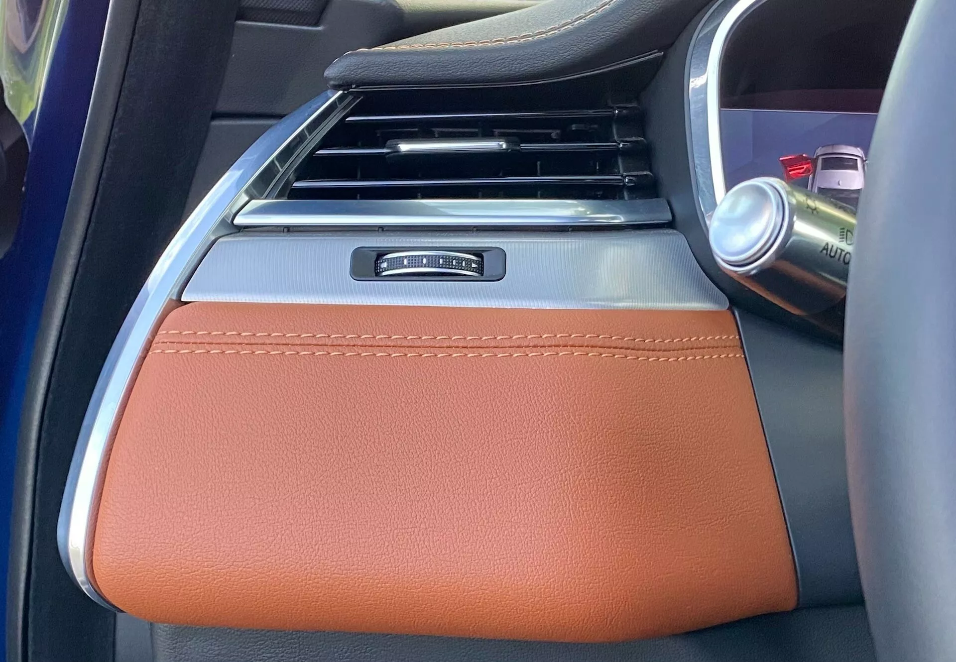 Dettaglio degli interni della Jaguar XF 2021, focalizzato sull'area del bracciolo centrale. L'immagine è scattata dal sedile posteriore guardando in avanti, mostrando un bracciolo in pelle marrone con cuciture visibili, che suggeriscono un livello di finitura di lusso. La parte superiore del bracciolo presenta un elemento in metallo, forse decorativo o funzionale. Dietro al bracciolo si vede la parte inferiore della console centrale con prese d'aria verticali, nere con accenti argento. A destra, un portabicchieri con una lattina indica spazio di archiviazione aggiuntivo. La parte argentata parzialmente visibile indica probabilmente un controllo per il riscaldamento, il raffreddamento o altre caratteristiche di comfort, mettendo in risalto il design interno moderno e premium.