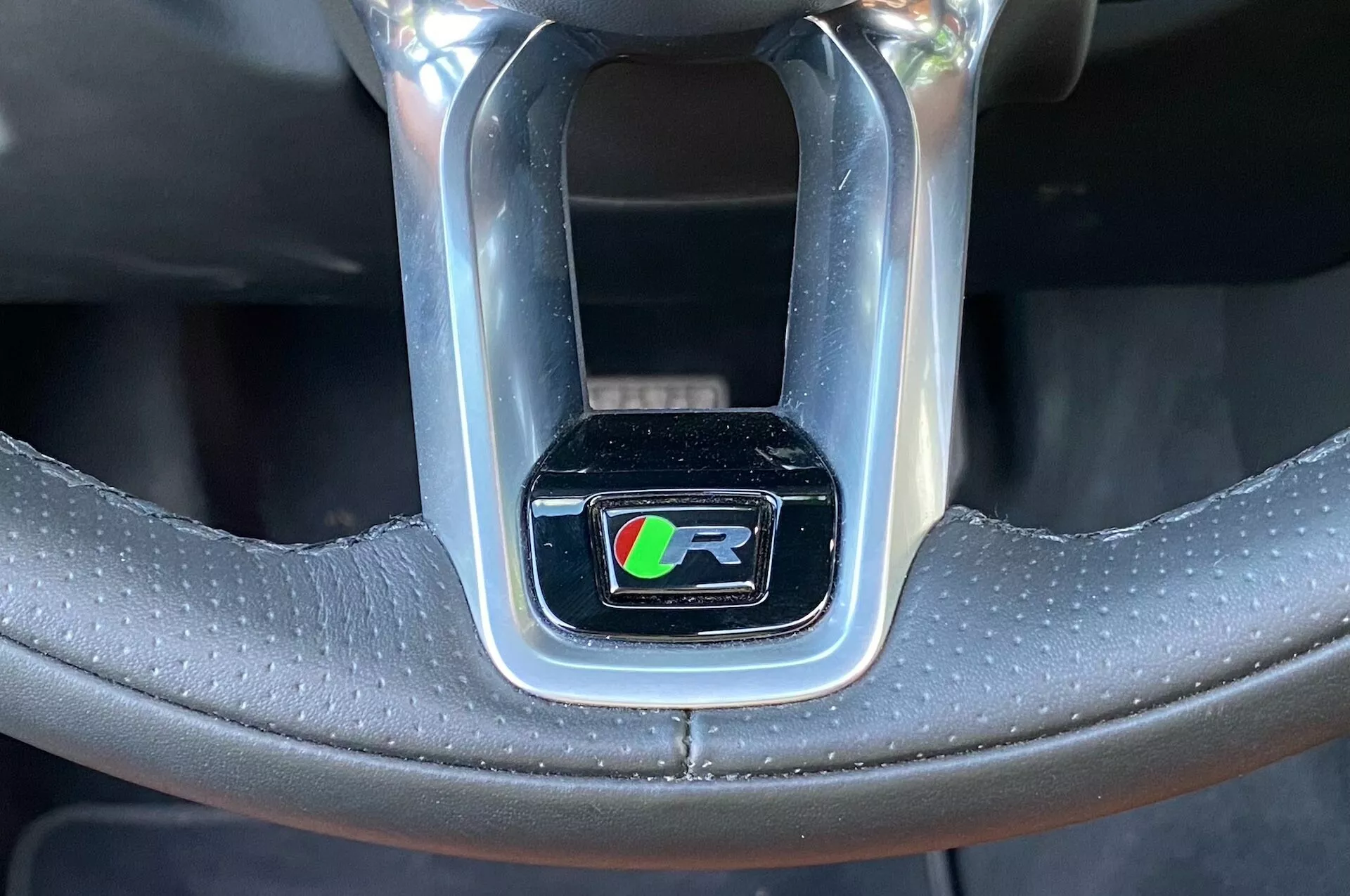 Primo piano di una sezione del volante di un'auto con il focus sul simbolo della linea di prestazioni ‘R’ racchiuso in un rettangolo cromato lucido. Il simbolo su sfondo nero presenta una ‘R’ stilizzata con strisce colorate in verde, blu, rosso e forse giallo. Il volante è realizzato in pelle nera, in parte perforata per una migliore presa e sensazione di lusso, con cuciture visibili. L'immagine ha un angolo frontale, leggermente verso l'alto, focalizzandosi esclusivamente sul simbolo, escludendo il resto degli interni dell'auto.