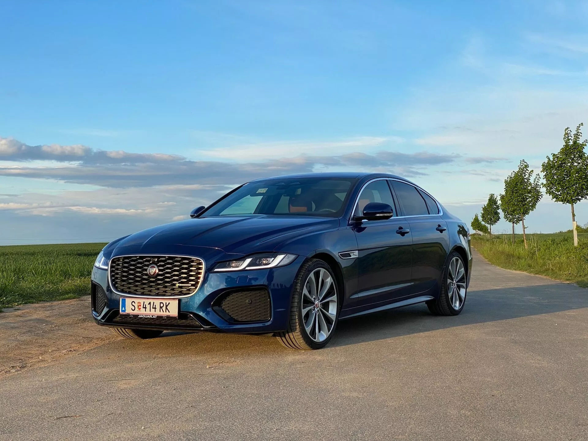 Jaguar XF 2021 blu parcheggiata su un'area pavimentata con un paesaggio naturale sullo sfondo, catturata da una vista frontale a tre quarti. Caratteristiche visibili includono griglia prominente con motivo a rete, fari LED angolari, luci fendinebbia integrate, cerchi in lega a più razze, linea del tetto elegante e pannelli laterali con linee di carattere ben definite, che mostrano elementi di design moderni e sportivi.