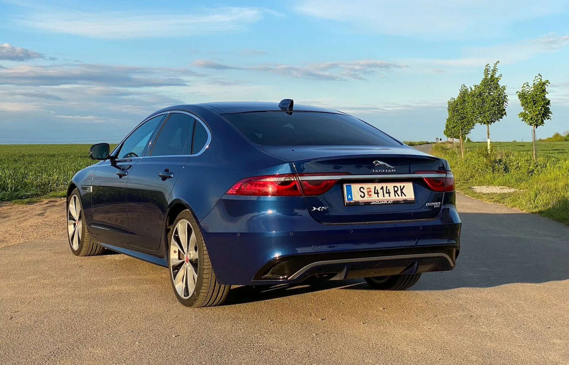 L'immagine mostra una vista di tre quarti posteriore di una Jaguar XF blue del 2021 parcheggiata su un'area non designata di ghiaia o terra a lato di una strada asfaltata, con il retro leggermente rivolto verso la telecamera. Le caratteristiche principali includono luci posteriori a LED orizzontali, baule con spoiler integrato, sensori di parcheggio sul paraurti posteriore, doppi terminali di scarico, cerchi in lega multirazze, antenna a pinna di squalo, specchietti laterali con indicatori di direzione integrati, finestrini chiusi con profili cromati e badging della modello. L'illuminazione suggerisce mattina o sera con luce morbida, vegetazione e cielo limpido sullo sfondo.