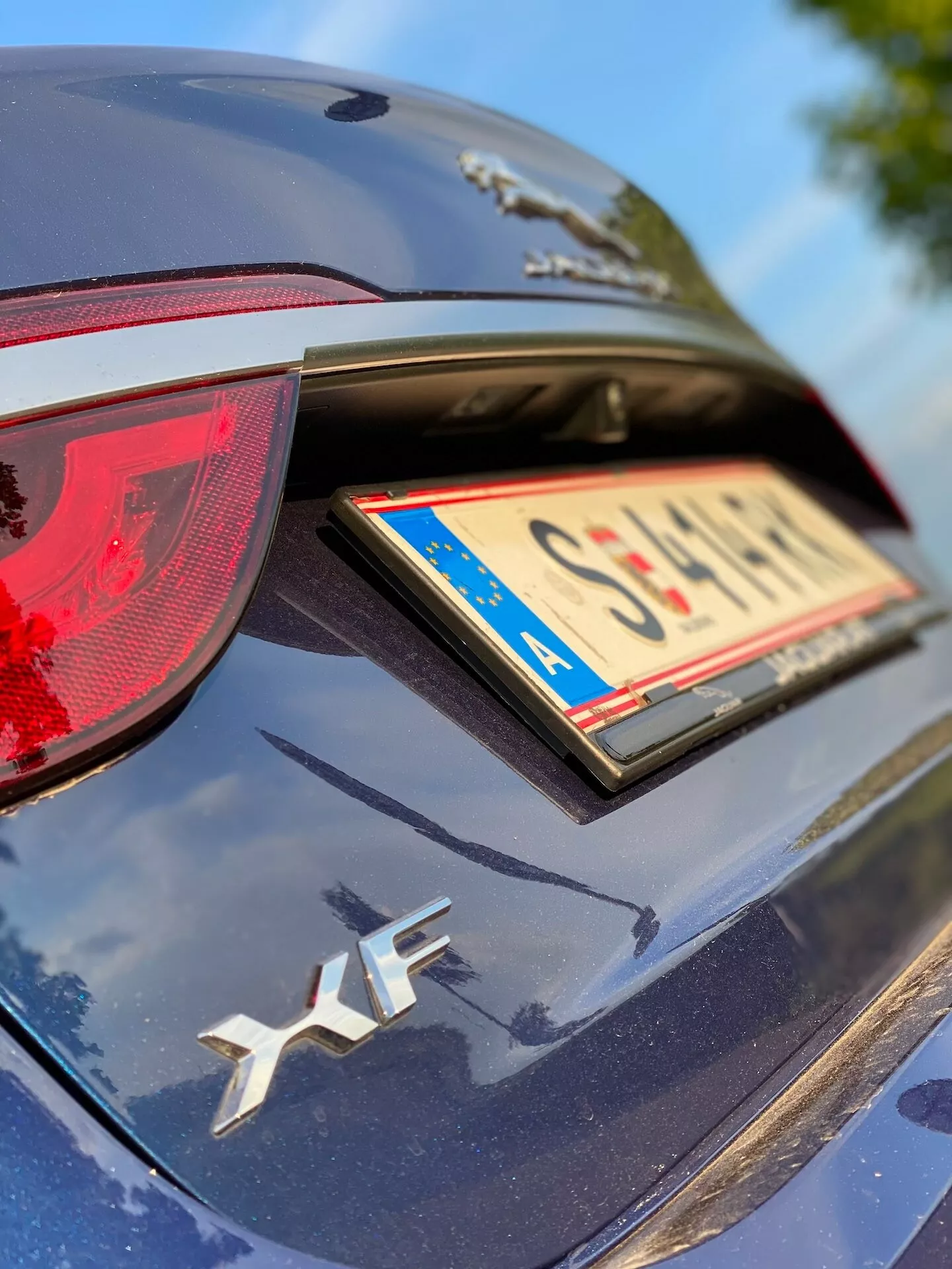 Primo piano della parte posteriore di un'auto di colore scuro, vista da un angolo obliquo alto. L'immagine si concentra sulla parte superiore destra della parte posteriore dell'auto, caratterizzata da un fanale posteriore dal design affilato sul lato destro, un coperchio del bagagliaio con un badge automobilistico 'XF', un portatarga con bordo a motivo di bandiera e un emblema parzialmente visibile sopra la targa. La vernice riflette la luce del sole, suggerendo che l'auto è ben tenuta o nuova. Complessivamente, le caratteristiche tecnologiche non sono distinguibili in questo scatto.