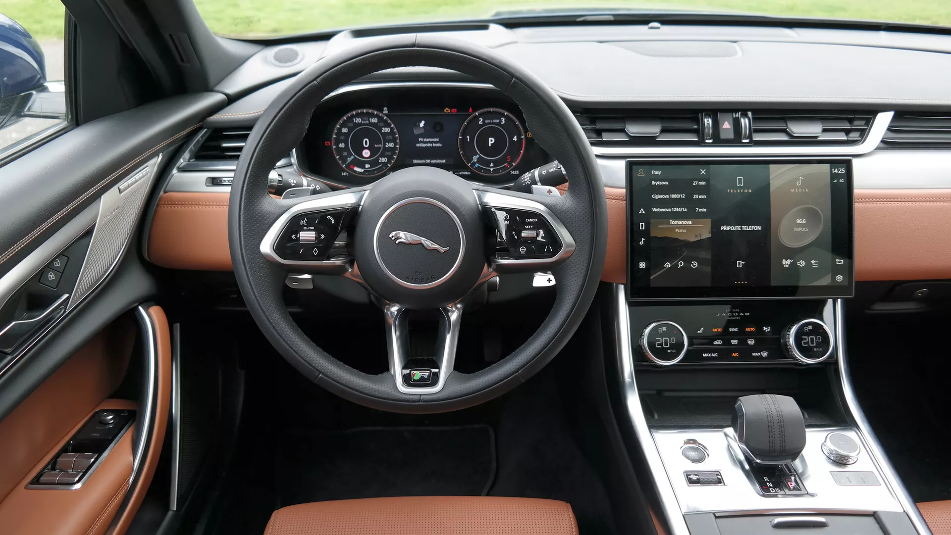 Scatto degli interni Jaguar XF 2021 focalizzato sull'area della cabina anteriore con un volante a tre razze e controlli multifunzione. Il quadro strumenti digitale visualizza le informazioni del veicolo. La console centrale ha un ampio sistema di infotainment touchscreen e un sistema di climatizzazione a doppia zona. Un selettore di marcia a rotore elettronico e un design interno premium a due toni nero e marrone/tan sono predominanti. Il pannello della porta e i sedili parzialmente visibili suggeriscono rivestimenti in pelle o simili e finestrini elettrici.