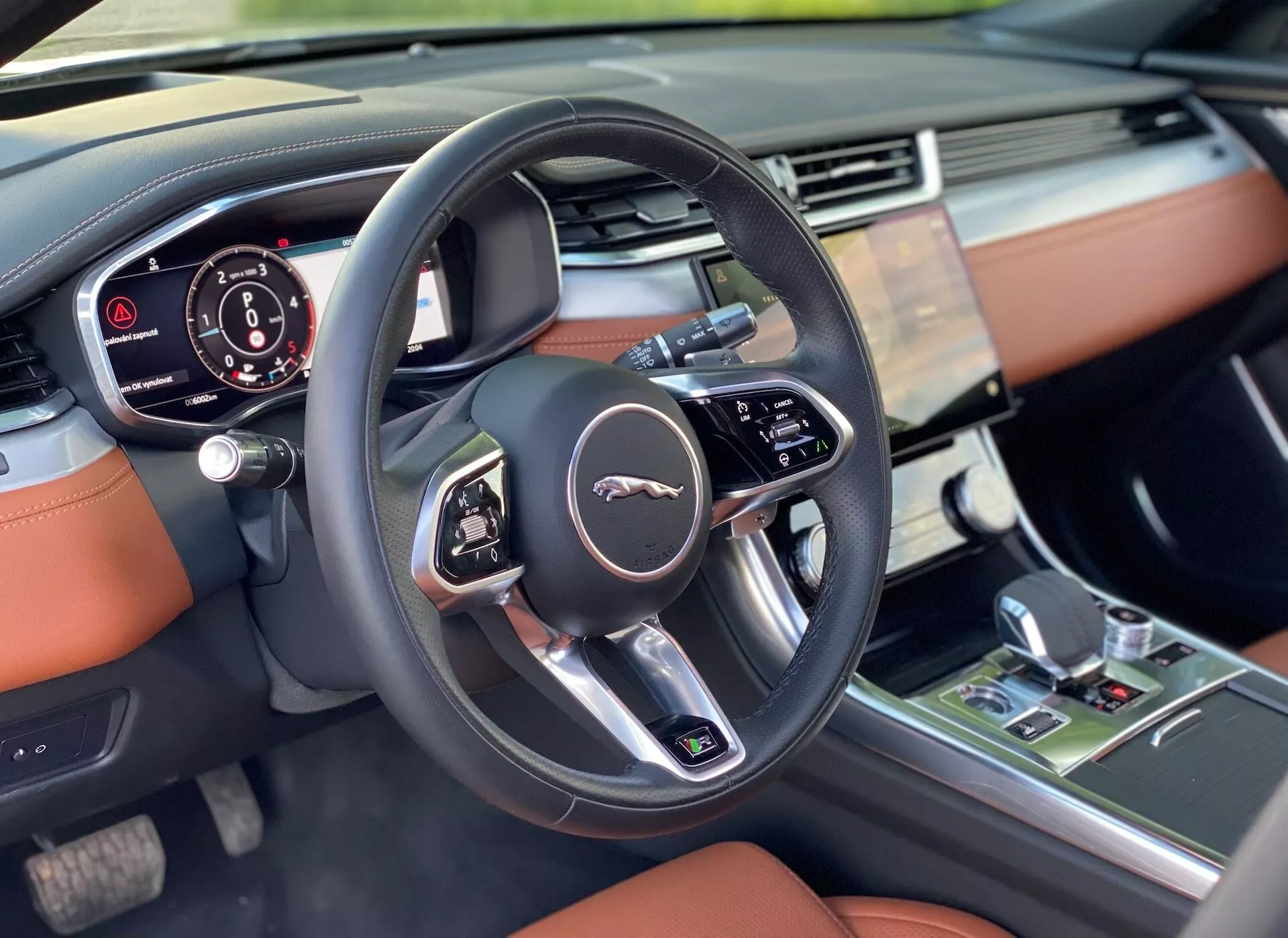 L'immagine mostra una vista ravvicinata dell'interno della Jaguar XF 2021 focalizzata sul lato del conducente dalla prospettiva del sedile anteriore del passeggero. In primo piano, il volante moderno a tre razze con pulsanti multifunzione e l'emblema Jaguar. Dietro al volante, il quadro strumenti parzialmente visibile mostra informazioni digitali. A destra si trova il sistema di infotainment dell'auto sulla parte centrale del cruscotto, probabilmente controllando l'audio, la navigazione e varie impostazioni. L'interno lussuoso presenta un cruscotto a tocco morbido con accenti in pelle cucita in un colore marrone scuro, toni più scuri, accenti metallici e una leva del cambio che suggerisce una trasmissione automatica e controlli per il clima e le modalità di guida. L'illuminazione di alta qualità mette in risalto i dettagli.