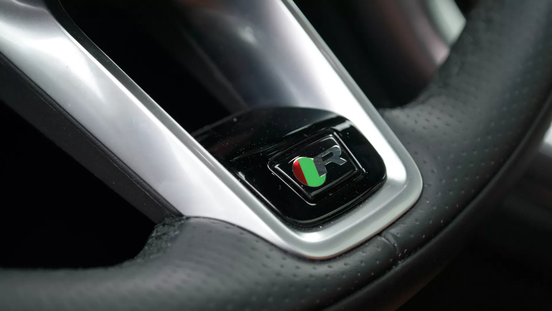 Primo piano della prospettiva del conducente del volante della Jaguar XF 2021, con display digitale al centro che mostra una bandiera a scacchi, suggerendo la modalità prestazioni attivata. L'immagine mette in evidenza il materiale in pelle di lusso con cuciture visibili e texture in fibra di carbonio, completato da un accento cromato intorno allo schermo, riflettendo una luce brillante.