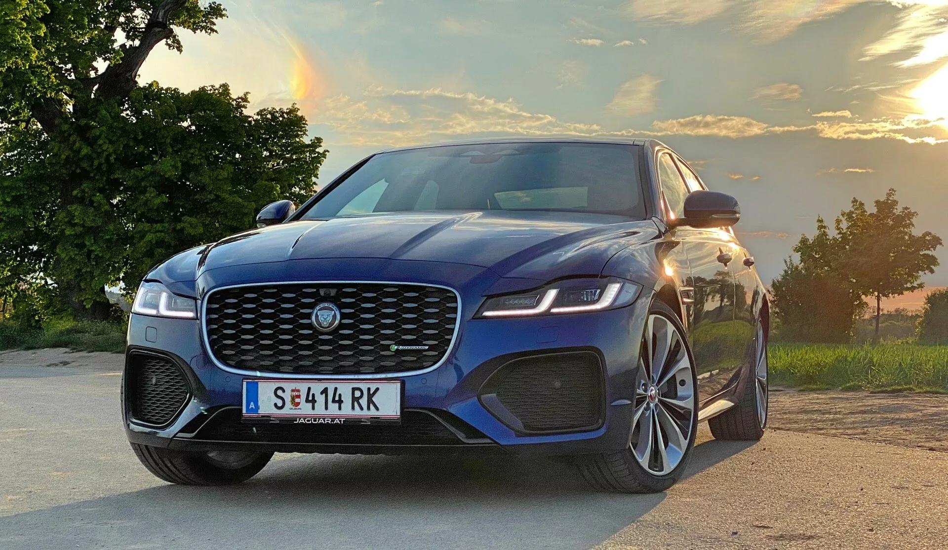 L'immagine mostra una Jaguar XF 2021 da un'angolazione bassa di tre quarti anteriore, mettendo in evidenza il suo design aerodinamico, la grande griglia con l'emblema del marchio, i fari a LED affilati, il paraurti pronunciato con ampie prese d'aria, le linee scolpite del cofano e un elegante profilo laterale. L'auto presenta grandi cerchi in lega multirazza, specchietti laterali con indicatori di direzione integrati, linee della carrozzeria distintive e finestrini leggermente oscurati. Situata su una strada o un'area di parcheggio con uno sfondo panoramico di alberi e un cielo al tramonto, il veicolo emana potenza e movimento in avanti.