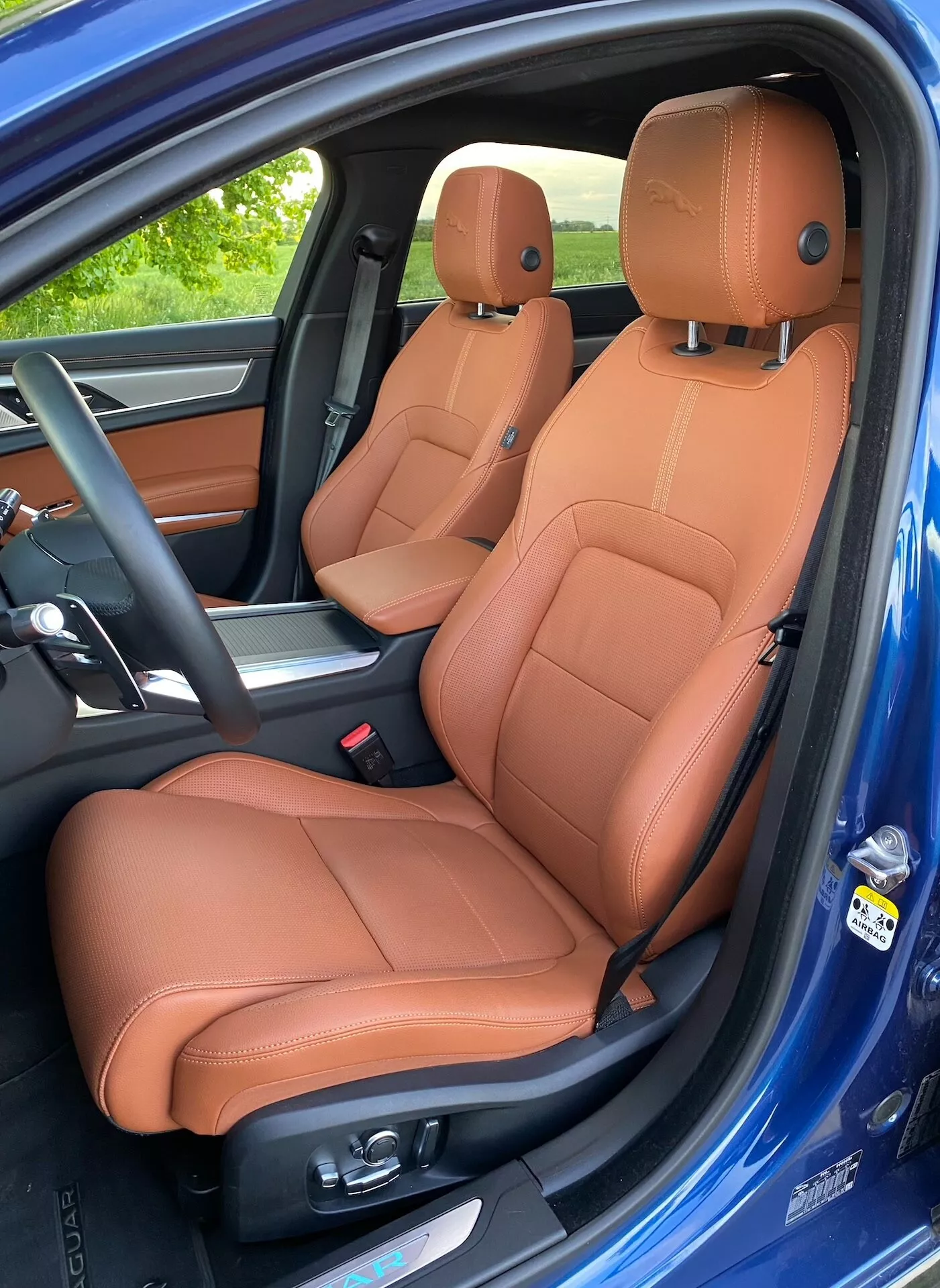 Vista angolata laterale dell'interno lussuoso di una Jaguar XF 2021, con particolare attenzione ai sedili anteriori rivestiti in pelle marrone con regolazioni elettroniche; console centrale parziale e etichetta airbag laterale visibili; vernice esterna blu vibrante che contrasta con lo schema bicolore dell'abitacolo.
