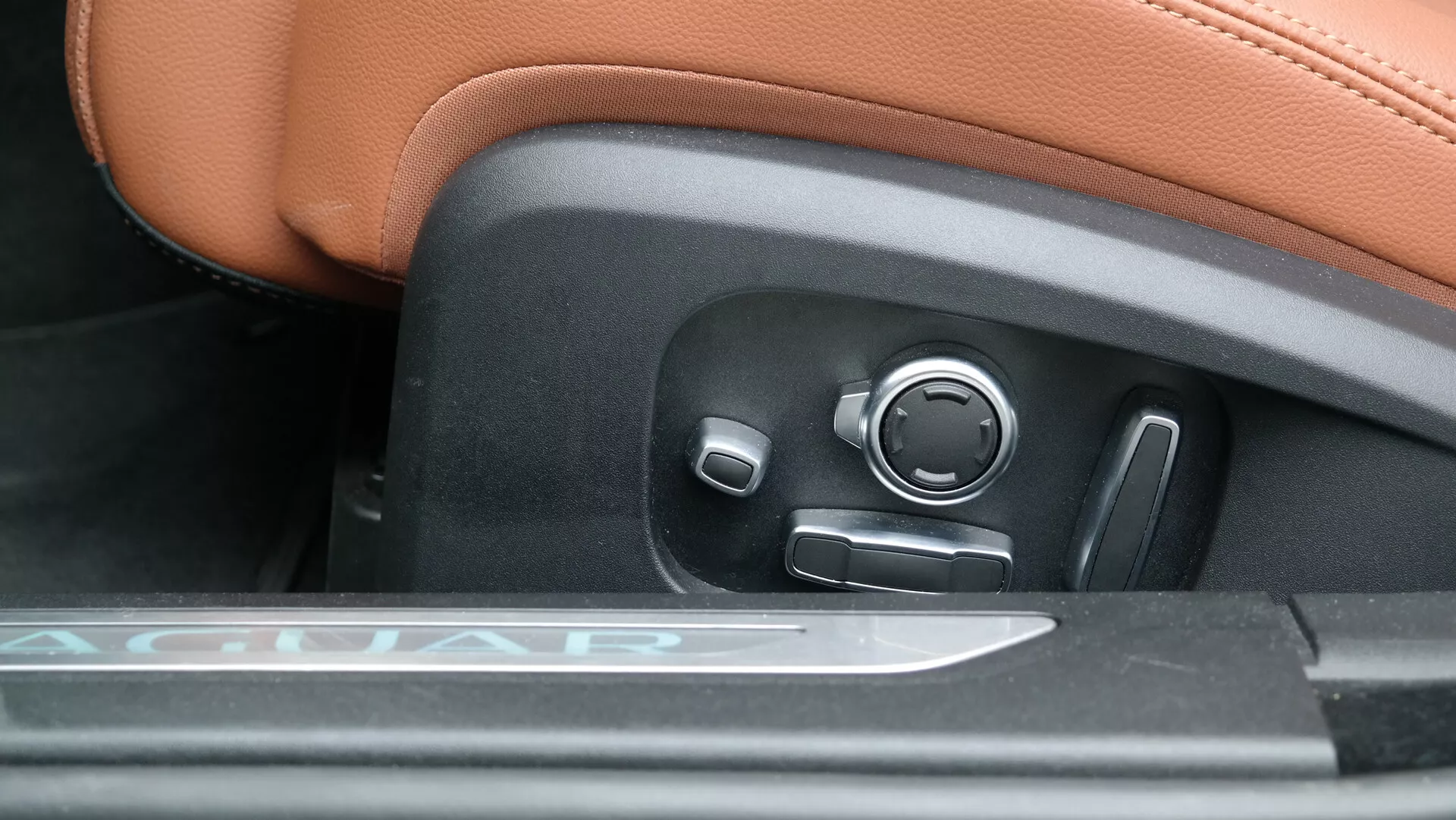 Dettaglio ravvicinato dei comandi di regolazione del sedile elettrico in una Jaguar XF 2021, con tappezzeria in pelle color cuoio con cuciture visibili, soglia d'ingresso in metallo con incisione 'JAGUAR' e avanzati comandi del sedile in plastica nera, tra cui una manopola a joystick con accento cromato, una leva per regolazioni di altezza e angolo e un controllo circolare per il supporto lombare.