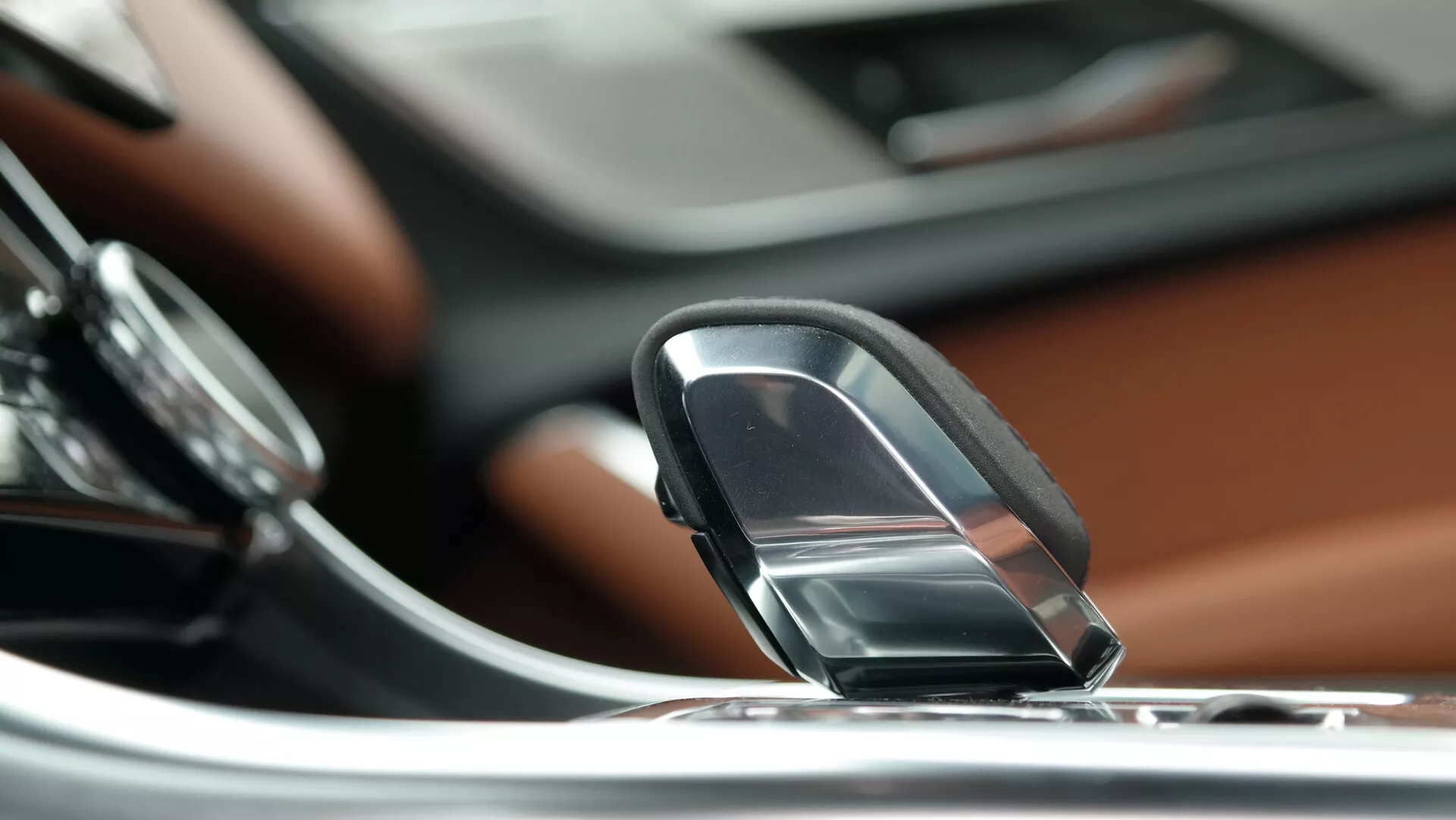 Vista ravvicinata dell'interno della Jaguar XF 2021, concentrata su un elegante joystick o selettore delle marce di tipo quadrante. La prospettiva è leggermente dall'alto e inclinata verso il basso, mettendo in risalto il selettore delle marce in primo piano. A sinistra è visibile parte di una manopola di controllo tonda relativa al sistema di infotainment o audio. Il cruscotto, rifinito con elegante legno, è visibile dietro il selettore delle marce, con uno schermo tattile integrato nella consolle centrale con accenti metallici sullo sfondo. Lo sfondo sfocato mette in risalto il selettore delle marce, suggerendo una profondità di campo ridotta. Ciò implica una trasmissione automatica e un sistema di infotainment moderno, indicando un veicolo di alta gamma e lusso.