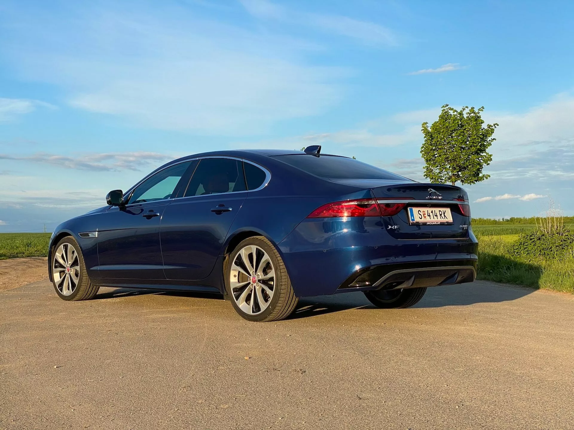 Moderna berlina a quattro porte in blu intenso vista da una prospettiva posteriore tre quarti su una strada di campagna. Il veicolo presenta un profilo aerodinamico ed elegante con un design fastback e cerchi in lega con design a più raggi. L'immagine mostra anche un tetto apribile, luci posteriori a LED moderne, una terza luce di stop e doppi tubi di scarico. Il cielo naturale e parzialmente nuvoloso sullo sfondo suggerisce un ambiente rurale aperto, aggiungendo una sensazione dinamica alla fotografia scattata da un angolo basso.