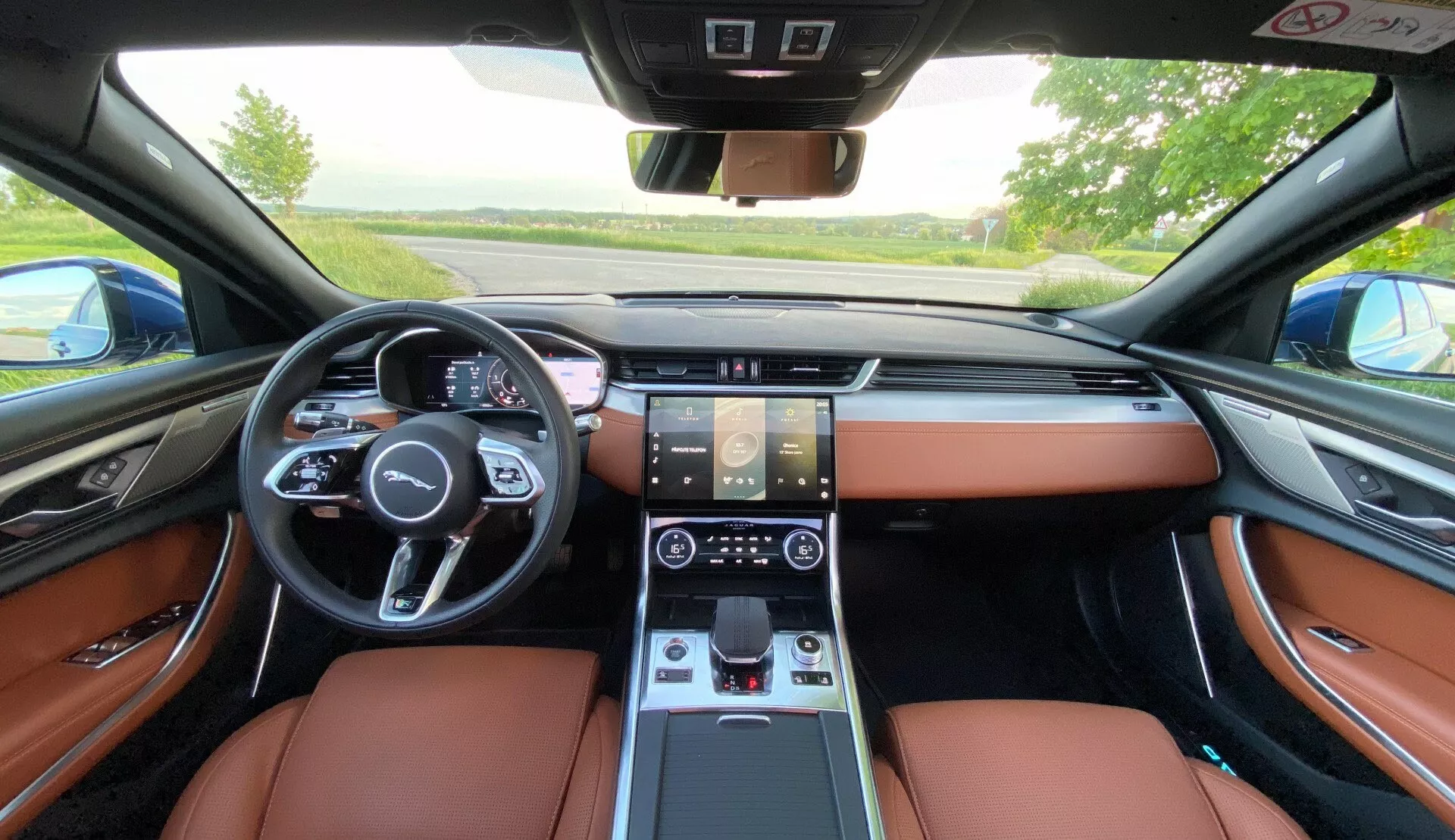 Vista degli interni del Jaguar XF 2021 dalla prospettiva del sedile posteriore, che mostra i lussuosi sedili in pelle marrone, il volante multifunzionale con comandi, il quadro strumenti digitale, il doppio display touch per infotainment e controllo del clima, il cambio elettronico e il design sofisticato del cruscotto con bocchette di ventilazione integrate.