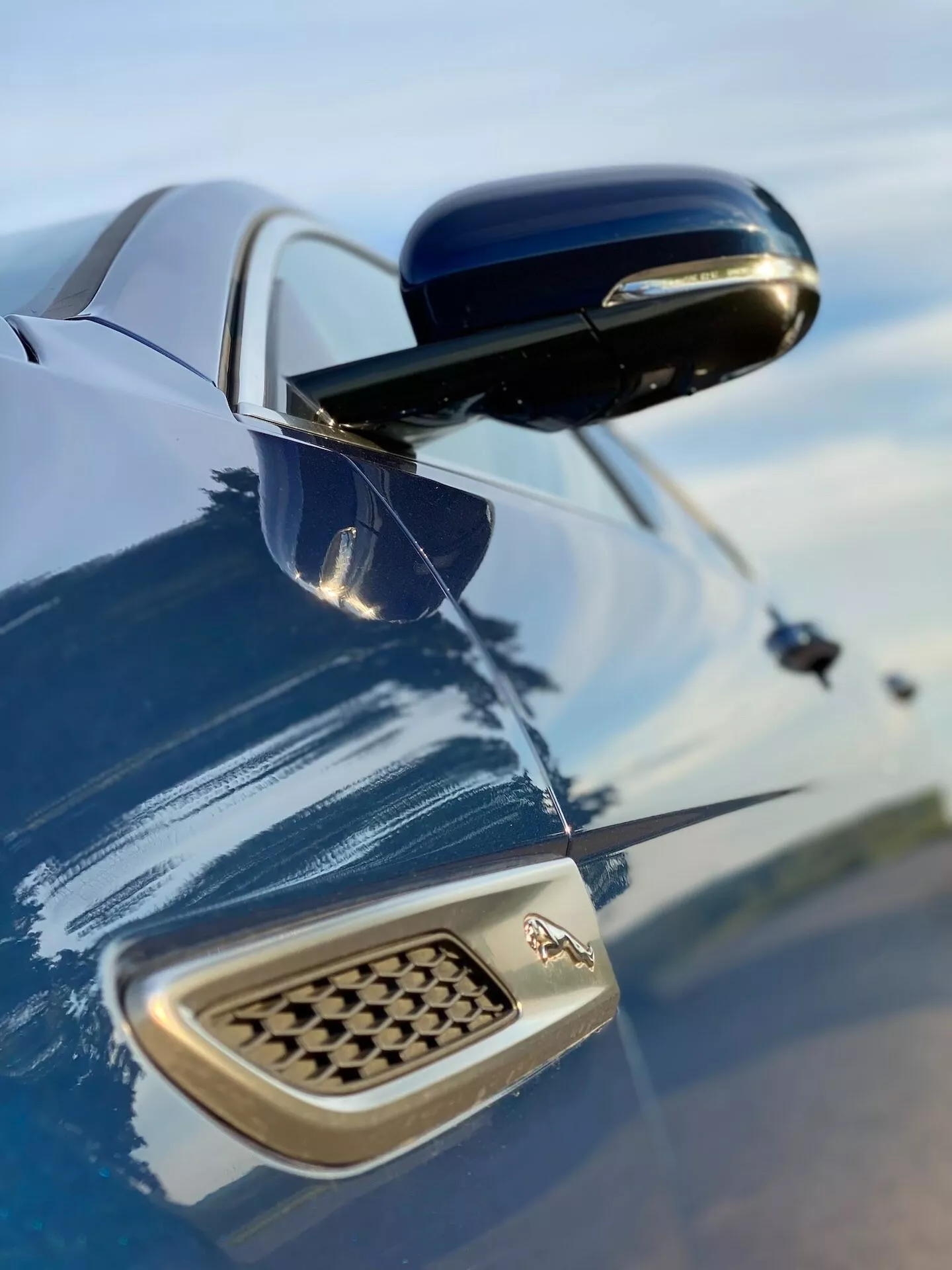 Vista ravvicinata del parafango laterale e dello specchietto laterale della Jaguar XF 2021 da un angolo basso verso l'alto, con vernice blu metallizzata profonda che riflette il cielo. Lo specchietto laterale prominente, con la sua carenatura nera lucida, contrasta con il colore della carrozzeria. Sull'ala laterale è visibile l'emblema del produttore e una presa d'aria a nido d'ape, che evidenziano elementi di design e prestazioni.