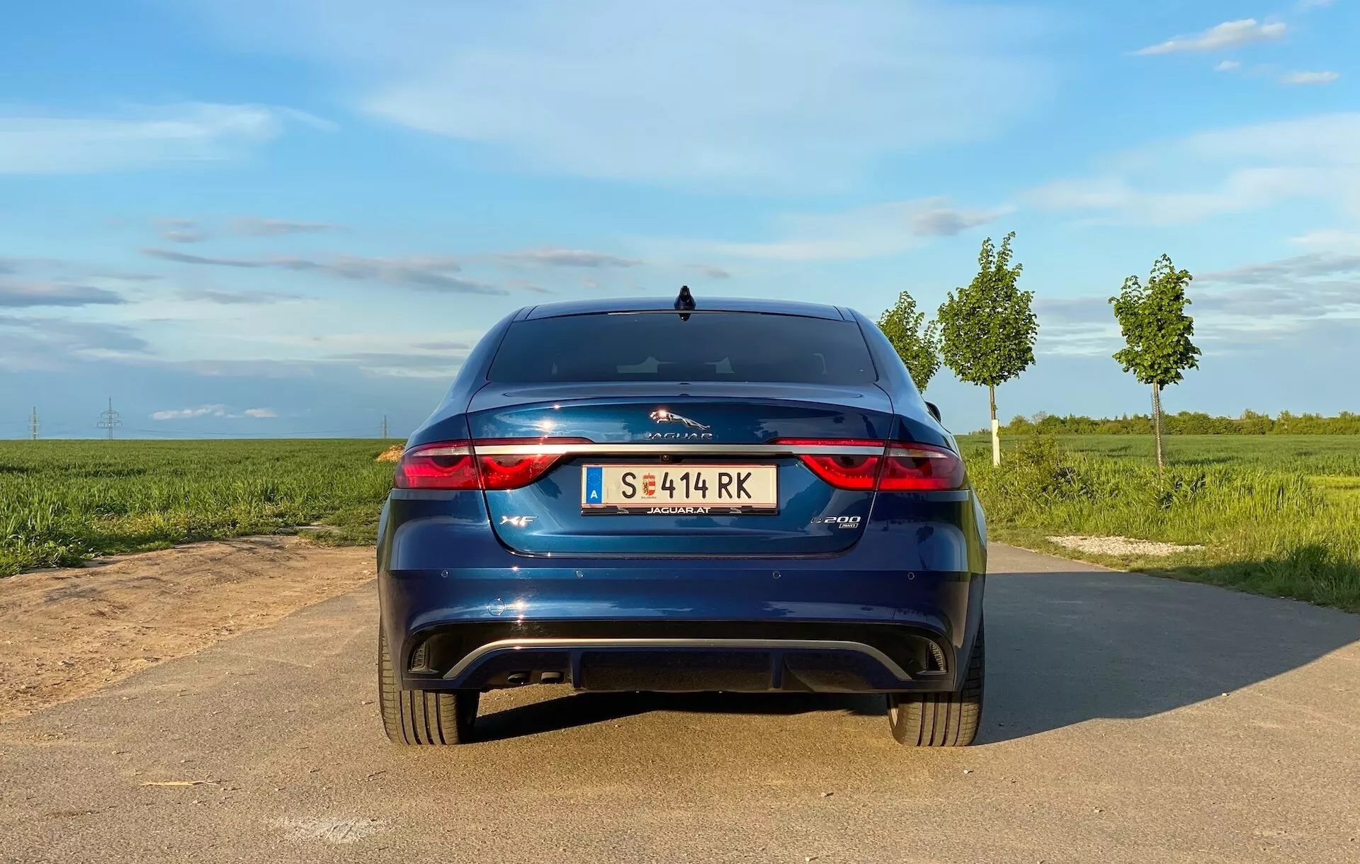 Vista posteriore di una Jaguar XF hatchback del 2021 su una strada asfaltata stretta, luce diurna con cielo sereno, campi aperti ai lati. Caratteristiche visibili: fanali posteriori a LED orizzontali, doppio scarico, logo centrale, emblema del modello a destra, targa centrale, piccolo spoiler posteriore, riflettori sul paraurti posteriore, possibili sensori di parcheggio/telecamera, antenna a pinna di squalo.