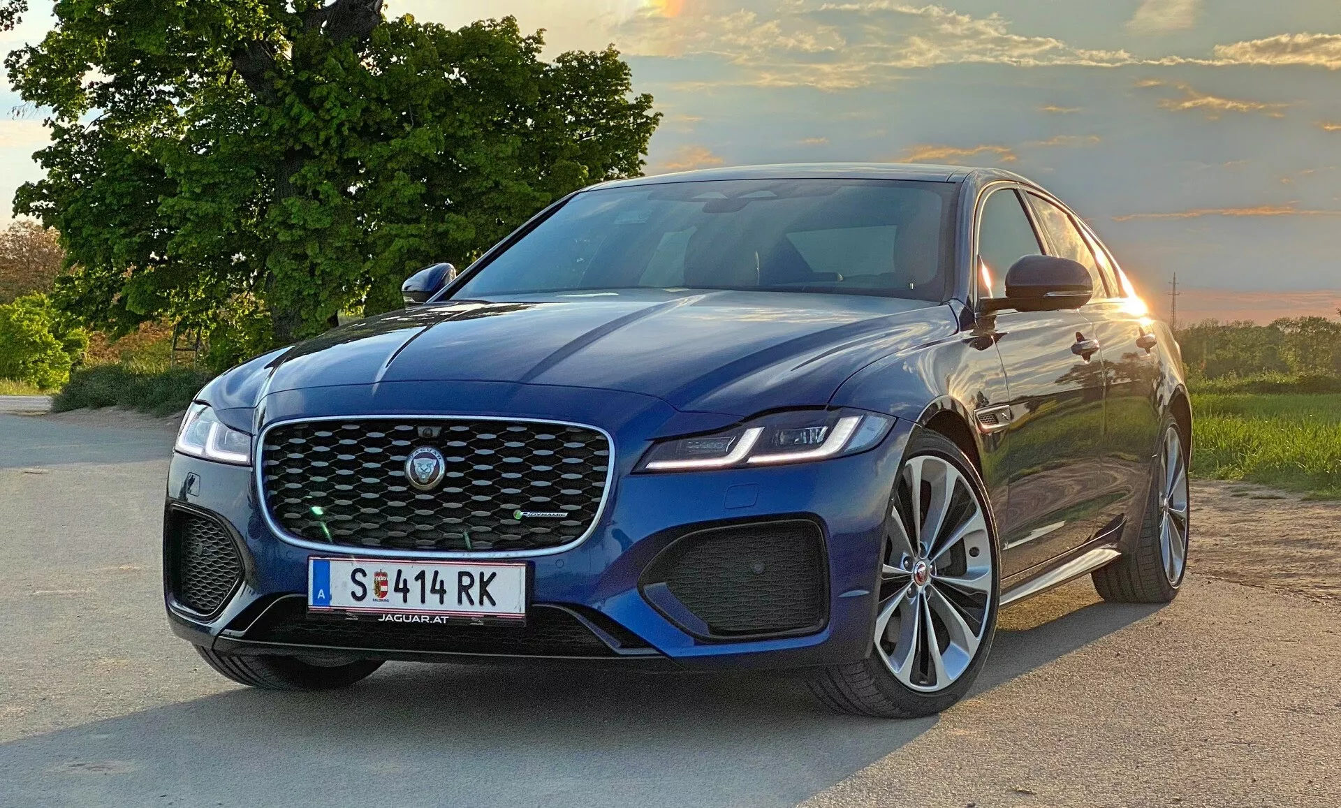 Berlina moderna Jaguar XF 2021 in vista frontale a tre quarti, catturando la grande griglia anteriore con l'emblema del produttore, i fari angolari eleganti, il paraurti anteriore sportivo con prese d'aria e parte del profilo laterale con linee di accento sollevate sul cofano, grandi ruote in lega e specchietti laterali con indicatori integrati. Il veicolo è posizionato su una strada con un paesaggio naturale che mostra alberi e un cielo al tramonto sullo sfondo.
