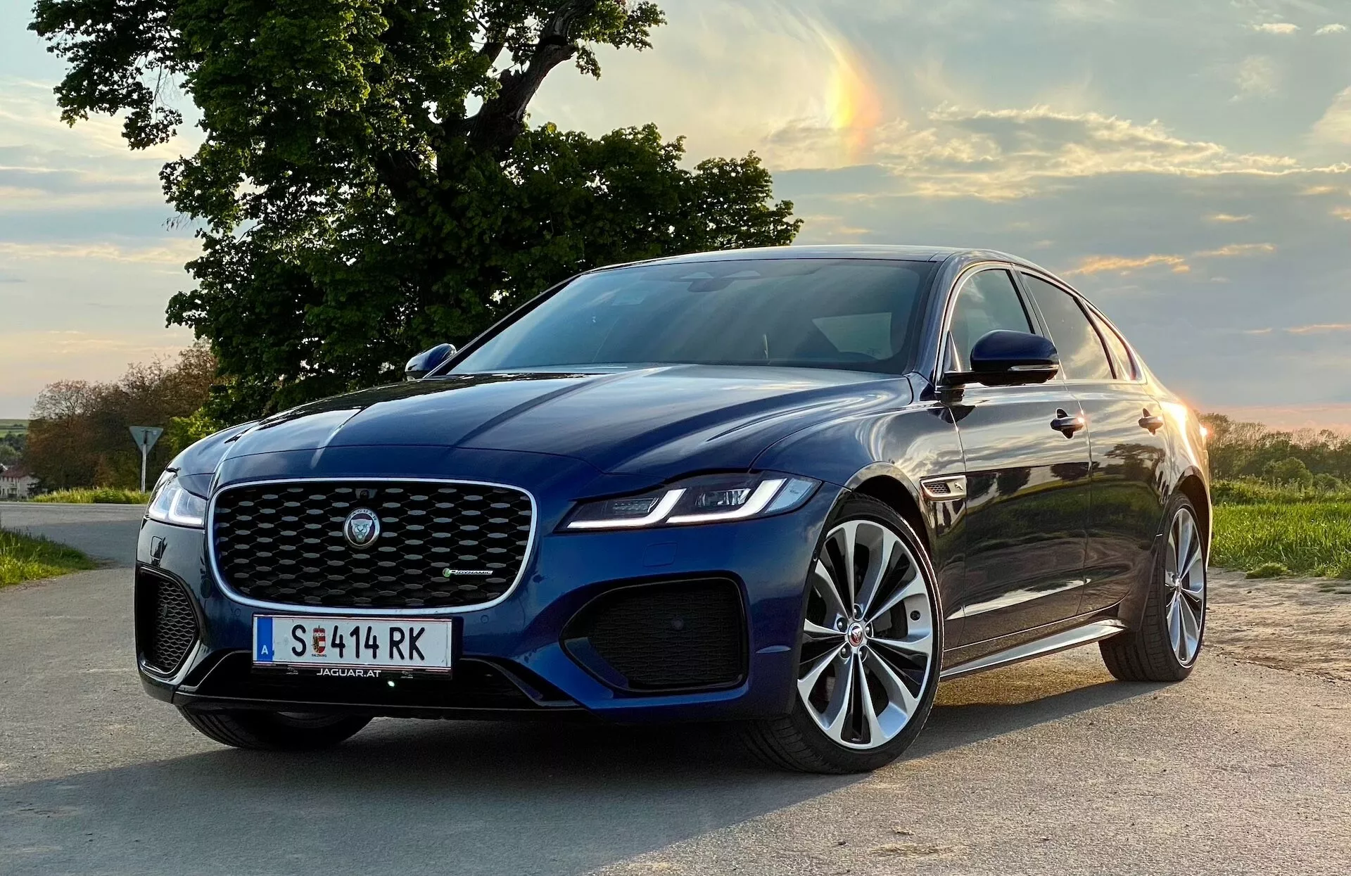 Sedan Jaguar XF 2021 ripreso da una angolazione frontale bassa a tre quarti, evidenziando la fascia anteriore, la griglia, il cofano e il profilo laterale del conducente. Il veicolo, di colore blu profondo con una superficie lucida, è parcheggiato su una strada asfaltata contro un paesaggio rurale con un sole al tramonto che proietta una luce calda. La griglia frontale prominente presenta un motivo distintivo, affiancata da eleganti fari LED. Il paraurti anteriore mostra prese d'aria e possibili sensori per i sistemi di assistenza alla guida, impreziosito da un basso spoiler. Le ruote in lega a più razze con pneumatici a profilo ribassato sottolineano il design orientato alle prestazioni della vettura.