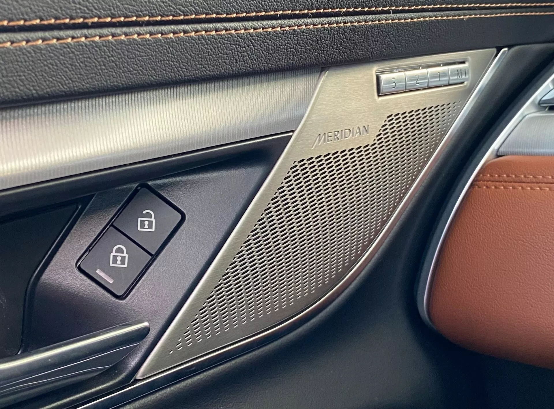 Vista ravvicinata del pannello della porta interna di una Jaguar XF 2021 dalla prospettiva di qualcuno seduto all'interno dell'auto. La parte superiore del pannello presenta una griglia dell'altoparlante con il marchio 'MERIDIAN', indicando un sistema audio di alta gamma di Meridian. Sotto l'altoparlante ci sono due pulsanti di controllo dei finestrini elettrici, che suggeriscono una funzionalità automatica a un tocco sia per i finestrini anteriori che posteriori, illuminati da una piccola luce LED. Il pannello della porta è realizzato in pelle marrone di alta qualità con dettagli di cuciture e plastica nera testurizzata intorno ai pulsanti. Gli accenti metallici intorno alla griglia dell'altoparlante e ai pulsanti accentuano l'aspetto lussuoso.