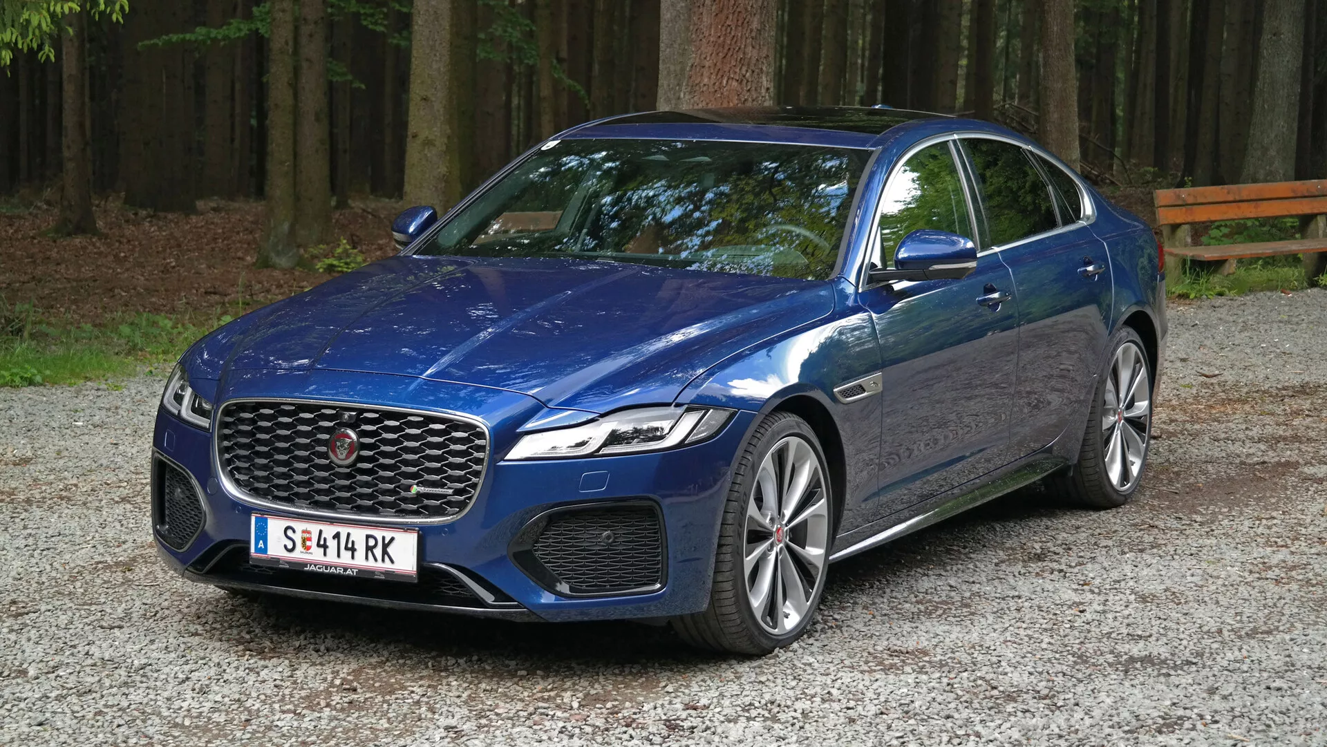 L'immagine ritrae una Jaguar XF 2021, una moderna berlina di lusso parcheggiata su una superficie ghiaiosa in un'area boschiva. Inquadrata da un'angolazione frontale a tre quarti, la foto mette in evidenza la griglia frontale prominente dell'auto con un emblema centrale, fiancheggiata da eleganti fari LED probabilmente dotati di luci diurne e forse di tecnologia adattiva. Sotto i fari, grandi prese d'aria possono servire a scopi sia estetici che funzionali. Il cofano presenta un design scolpito con creste rialzate, e il parabrezza inclinato suggerisce un'efficienza aerodinamica. Il profilo laterale rivela grandi cerchi in lega con un design multirazza e pneumatici a basso profilo, che possono indicare capacità prestazionali. Una linea di carattere corre lungo il lato, aggiungendo una sensazione di movimento, mentre gli specchietti retrovisori in tinta carrozzeria con indicatori di direzione integrati e le finiture cromate delle finestre accentuano l'aspetto di lusso del veicolo.
