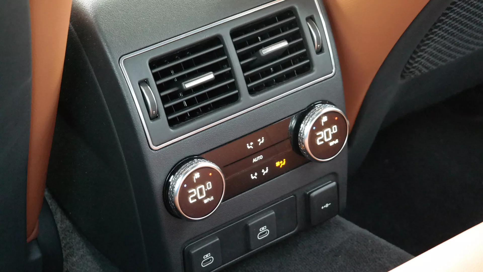Vista dettagliata della console centrale del controllo del clima del passeggero posteriore in un veicolo moderno, probabilmente la Jaguar XF 2021, che mostra le manopole di controllo del clima a doppia zona con display digitali impostati a 20 gradi, circondate da accenti neri e metallici. Include pulsanti per funzioni climatiche aggiuntive e due bocchette d'aria rettangolari sopra i controlli.