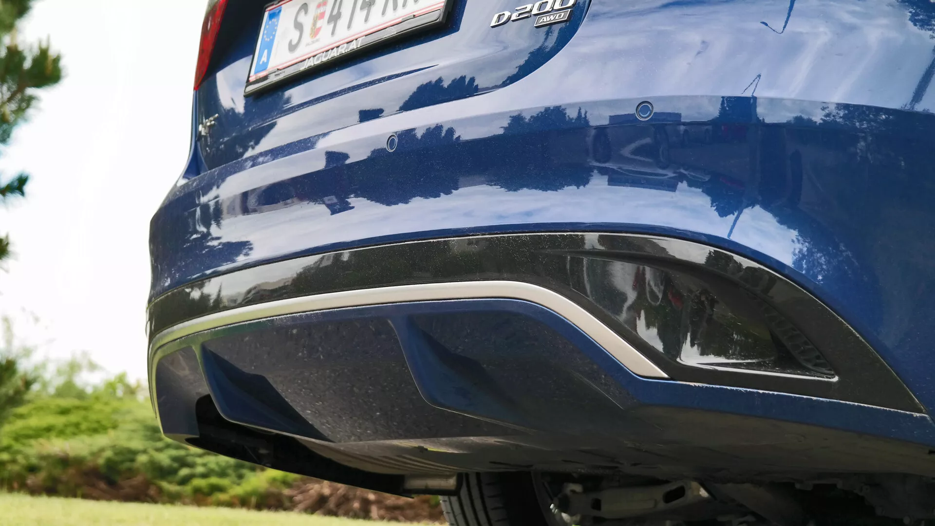 Dettaglio ravvicinato della sezione posteriore di una Jaguar XF 2021 blu, ripresa da un'angolazione obliqua a livello del suolo, che mostra il paraurti integrato tipo diffusore nero e grigio argento, la vernice blu metallizzata con riflessi ambientali e il badge 'DDDI' sul lato sinistro del coperchio del bagagliaio, senza luci posteriori o tubi di scarico visibili.
