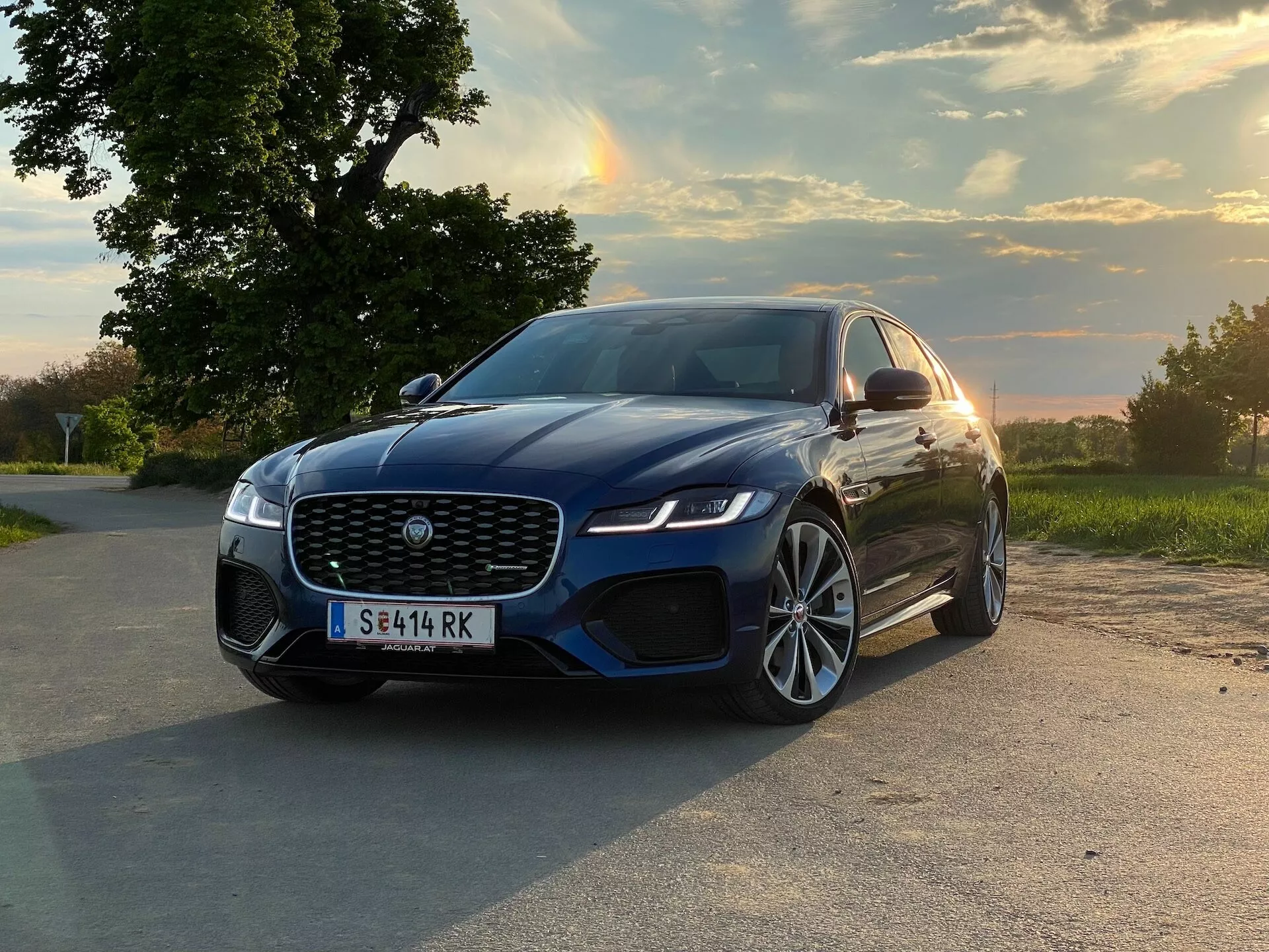 Fotografia di una berlina di lusso moderna, specificamente una Jaguar XF 2021, ripresa da un angolo basso e ravvicinato leggermente spostato verso il lato passeggero. L'auto è posizionata su una strada asfaltata in un ambiente rurale o suburbano con uno scenario naturale sullo sfondo, che suggerisce mattina presto o tardo pomeriggio in base ai toni caldi e all'angolo basso della luce solare che si riflette sulla carrozzeria. Il veicolo presenta una grande griglia frontale con un emblema prominente, fari LED eleganti, un sensore di parcheggio anteriore visibile, un cofano scolpito, grandi ruote in lega, vernice blu metallica profonda, specchietti laterali aerodinamici e un potenziale sistema di assistenza al parcheggio.