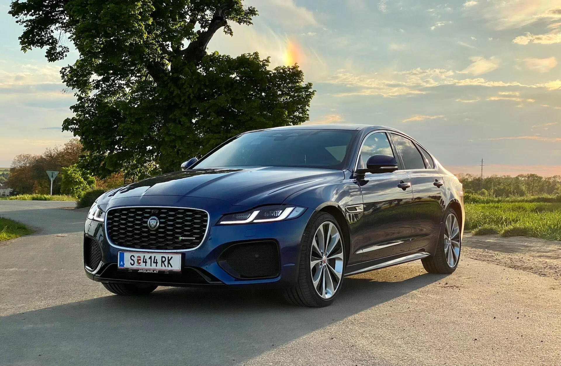 Questa immagine ritrae la Jaguar XF 2021, una berlina di lusso, posizionata in diagonale su una strada rurale. L'auto è leggermente orientata verso destra, mostrando il quarto anteriore sinistro incluso la grande griglia a rete, i fari LED eleganti, il diffusore d'aria integrato con sensori di parcheggio, gli specchietti laterali montati sulle porte, probabilmente con indicatori di direzione, i cerchi in lega multi-razze con pneumatici a profilo basso e le linee aerodinamiche della carrozzeria con profilo slanciato. La scena è ambientata in un ambiente rurale sereno con calda illuminazione naturale serale, evidenziando il design premium e le caratteristiche tecnologiche avanzate della vettura.
