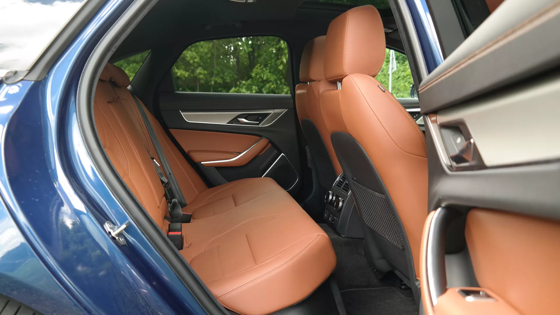 Immagine che mostra l'interno posteriore di una Jaguar XF 2021, vista dall'esterno attraverso la porta posteriore aperta. Le caratteristiche includono sedili in pelle marrone caldo, sedile del passeggero anteriore visibile con cuciture dettagliate, artigianato di qualità, design ergonomico per il comfort e pannello porta con comandi per l'intrattenimento in auto.