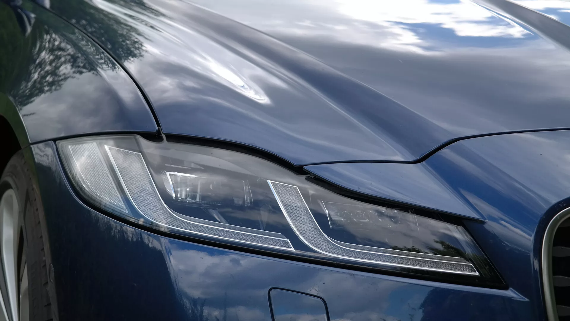 Primo piano del quarto anteriore sinistro di una Jaguar XF 2021 incentrato sull'insieme dei fari a LED angolari, caratterizzato da un design moderno con strisce e punti orizzontali, cofano scuro lucido e linee aerodinamiche, esemplificando lusso e tecnologia automobilistica avanzata.