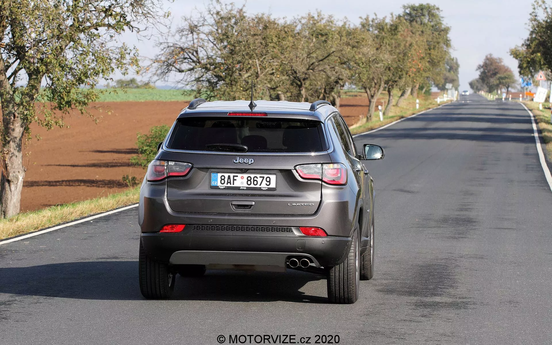Vista posteriore di una Jeep Compass 2017 che si allontana su una strada a due corsie, mostrando elementi di design moderni come antenna a pinna di squalo, tergicristallo posteriore, luci posteriori a LED, sensori di parcheggio integrati, doppi tubi di scarico e targa in stile europeo con logo Jeep chiaramente visibile. Il veicolo sembra essere in movimento, catturando il funzionamento reale con potenziali tecnologie di assistenza alla guida.