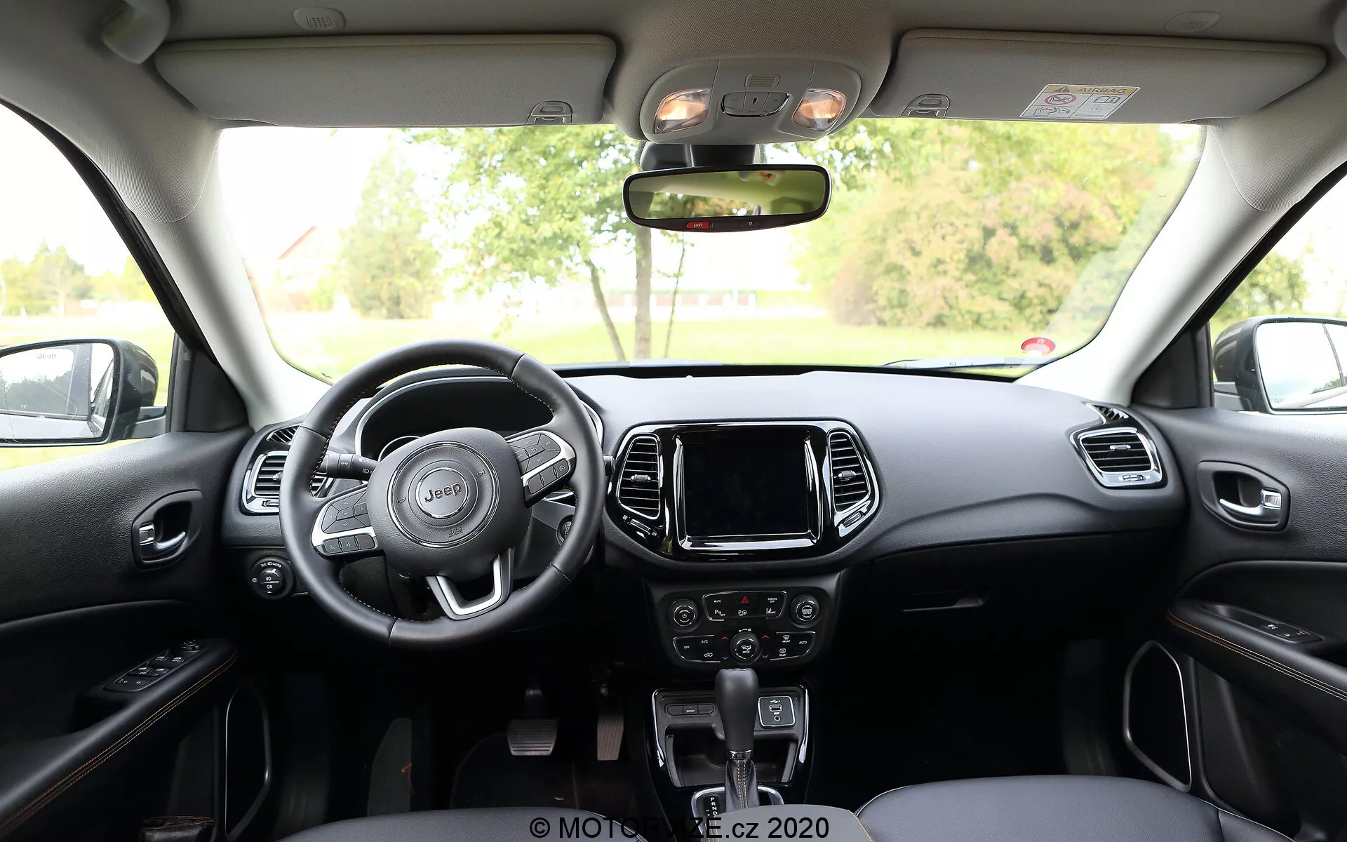 Vista interna della Jeep Compass 2017 dal sedile posteriore, che mostra un volante a tre razze con comandi integrati, console centrale con sistema di infotainment touchscreen, comandi manuali per audio e clima sotto di essa e leva del cambio automatico. Il cruscotto e i sedili anteriori presentano uno schema bicolore con accenti metallici o plastici, mentre sopra ci sono i comandi per l'illuminazione interna e il tetto apribile.