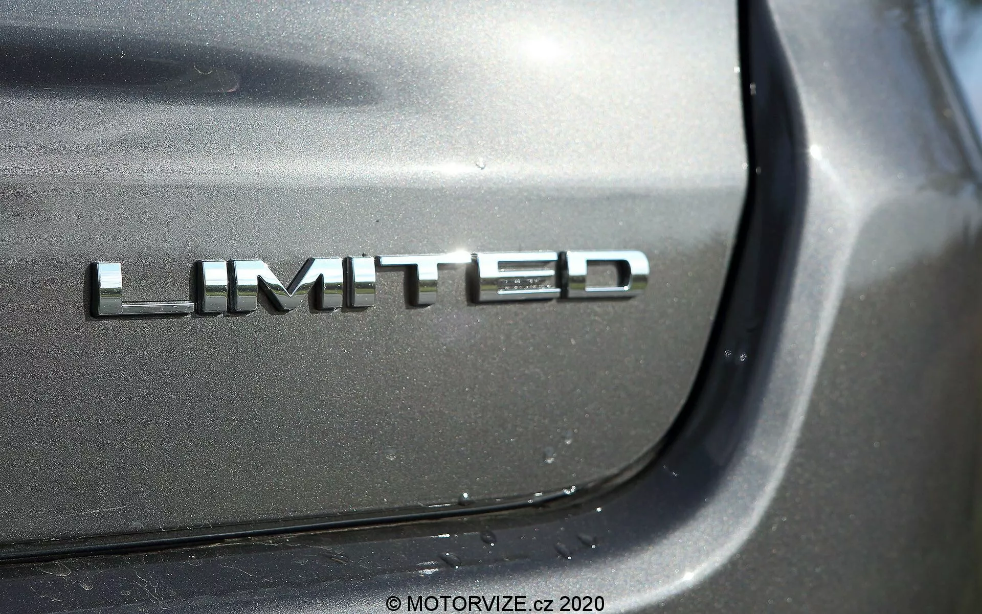 Dettaglio ravvicinato del badge Limited della Jeep Compass 2017 con finitura cromata sul cofano posteriore; la fotografia si concentra sul badge da una prospettiva angolata con un riflesso luccicante sotto la luce diretta, sfocando i dettagli circostanti dell'auto per enfatizzare la verniciatura metallica di alta qualità.