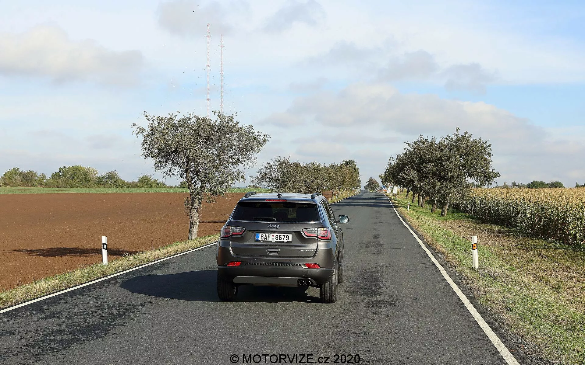 L'immagine mostra la vista posteriore di una Jeep Compass 2017 che percorre una strada di campagna a due corsie in una giornata di sole con nuvole sparse. Il veicolo, posizionato sul lato destro della corsia, mostra il portellone, il paraurti, le luci posteriori, il lunotto, la targa, parte del tetto con l'antenna a pinna di squalo e la parte superiore degli pneumatici posteriori. L'auto occupa una parte significativa del frame, consentendo una chiara visione dei dettagli delle sue caratteristiche. La strada si curva dolcemente a destra in lontananza, circondata da campi, alberi e pali stradali. Le luci dei freni dell'auto non sono accese, suggerendo una guida normale.