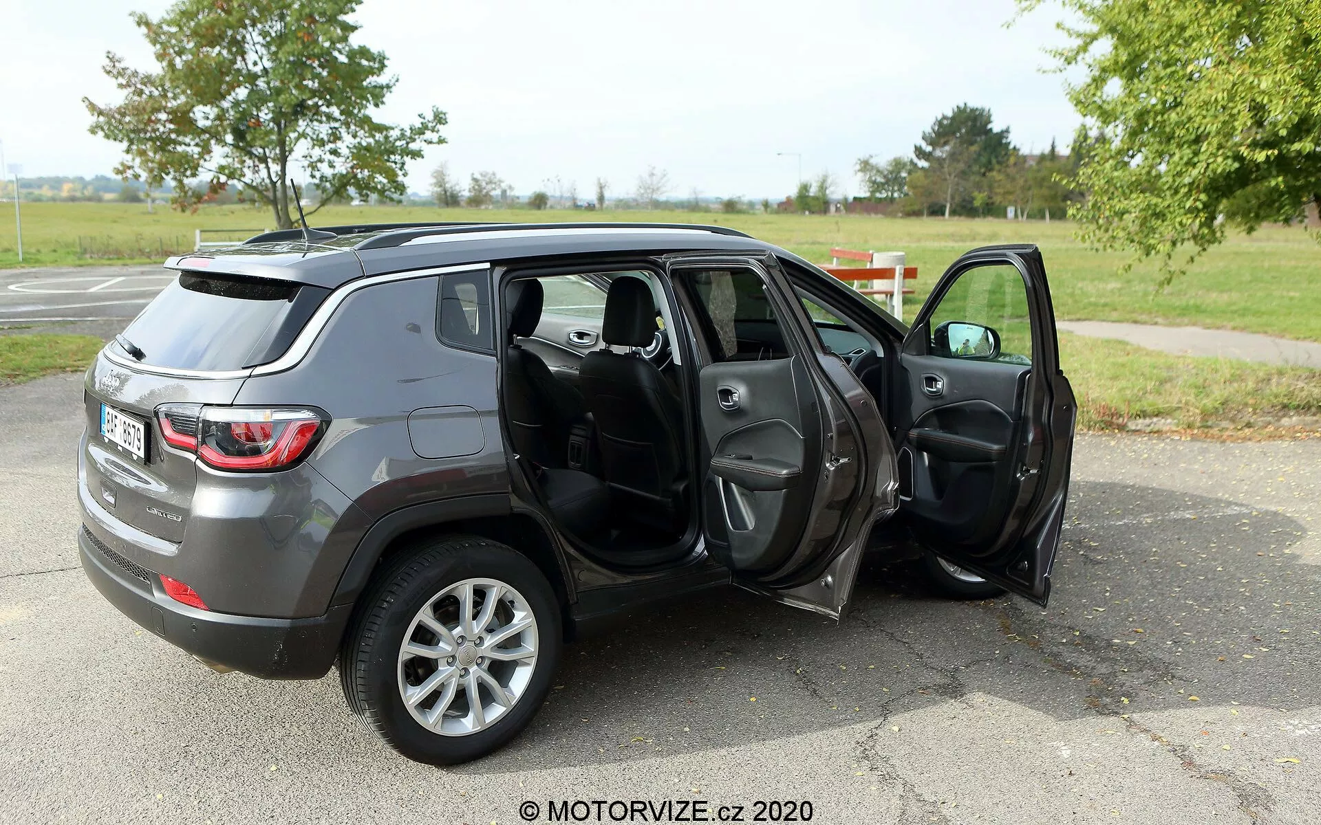 Immagine di un SUV di medie dimensioni Jeep Compass 2017 parcheggiato su una superficie asfaltata in un ambiente simile a un parco con erba e alberi sullo sfondo. La foto è scattata da un angolo posteriore di tre quarti, mostrando il profilo e la parte posteriore del veicolo. Entrambe le portiere del lato passeggero sono aperte, rivelando l'interno in tessuto scuro o pelle, inclusi i sedili anteriori e posteriori, con possibili caratteristiche come i finestrini e gli specchietti elettrici. Il SUV ha una finitura metallica grigia ed è dotato di cerchi in lega a cinque razze, il che indica un livello di allestimento più alto o un upgrade opzionale delle ruote. La filigrana 'MOTORVIZE.cz 2020' suggerisce che l'immagine sia di una recensione di auto o di un sito web automobilistico.