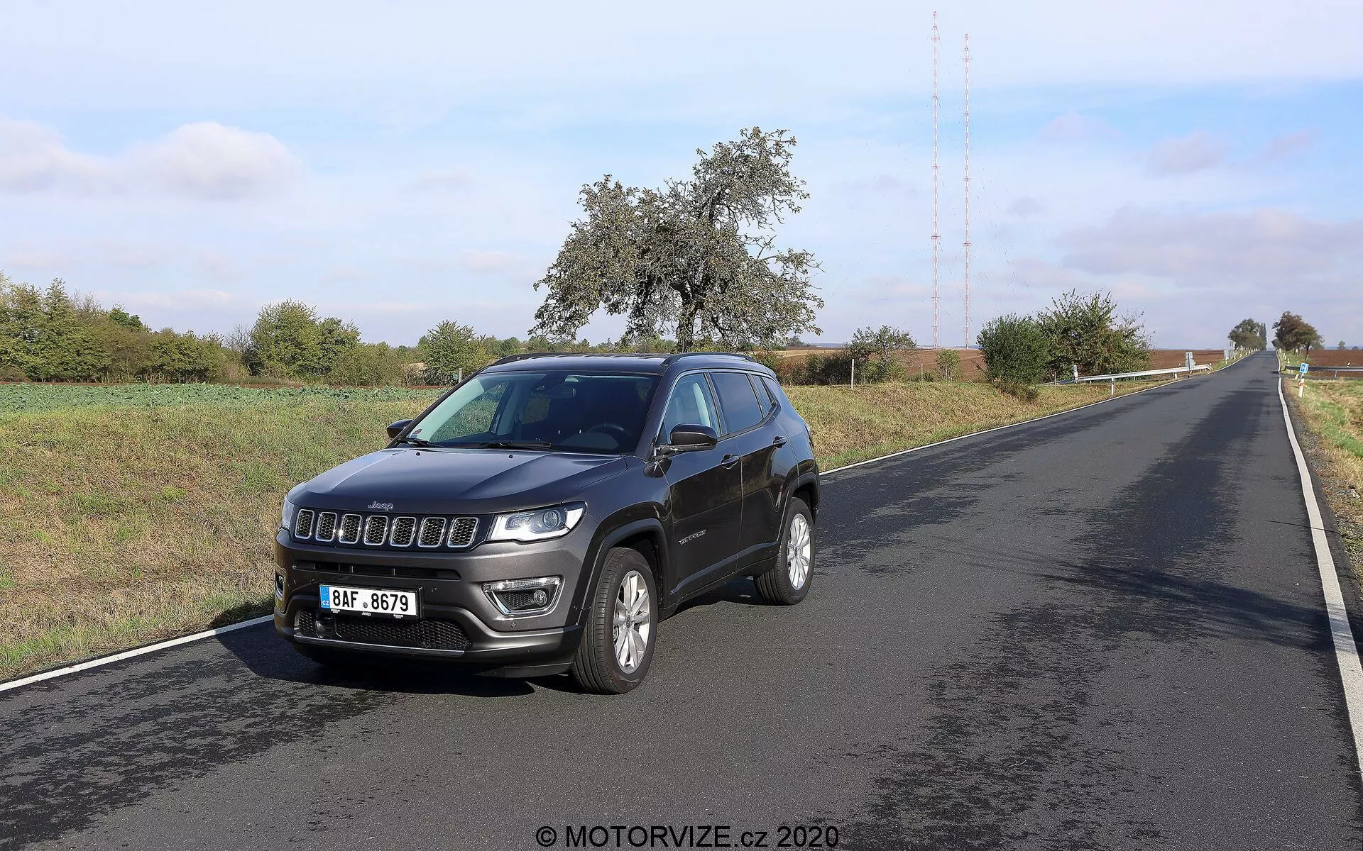 Immagine di una Jeep Compass 2017 fotografata da un'angolazione frontale di tre quarti. Il veicolo, posizionato sul lato destro di una strada a due corsie, mostra il suo profilo anteriore e laterale. Le caratteristiche principali includono una griglia moderna, fari prominenti con luci diurne a LED, cerchi in lega, indicatori di direzione integrati sugli specchietti laterali, un'antenna sul tetto e fari fendinebbia sul paraurti anteriore. L'ambiente sembra rurale con cielo sereno e vegetazione lungo la strada.