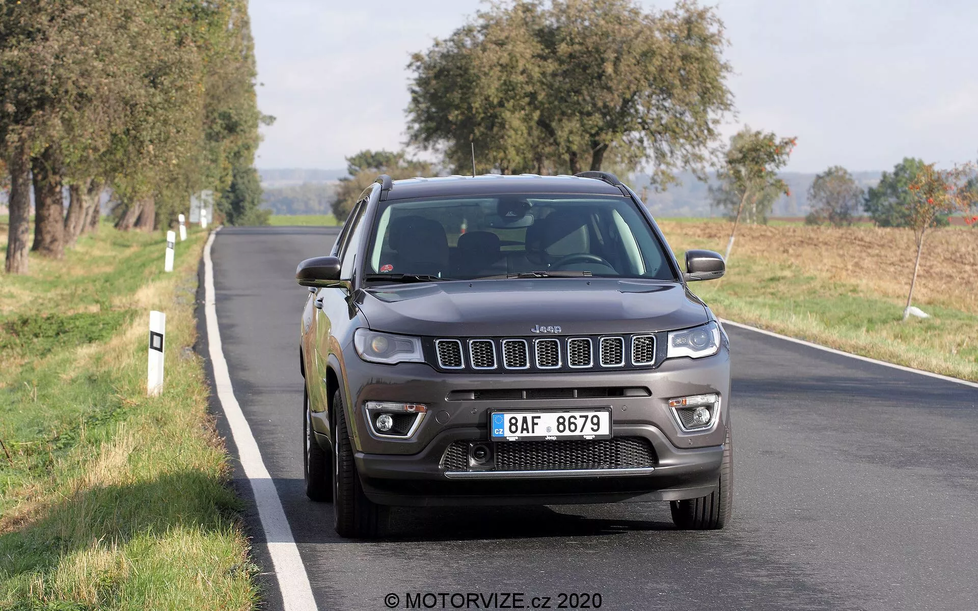 Immagine di una Jeep Compass del 2017 che guida su una strada a due corsie, fotografata da un angolo frontale di tre quarti. L'iconica griglia a sette slot del veicolo, i fari dettagliati con luci diurne a LED, i fendinebbia e le ruote in lega multirazze sono chiaramente visibili. Il colore grigio scuro o nero della Jeep le conferisce un aspetto solido e robusto, con la luce naturale del giorno che mette in risalto le sue caratteristiche. Il veicolo sembra essere in movimento, con una leggera sfocatura sulle ruote. Gli alberi e i campi aperti sullo sfondo suggeriscono un ambiente rurale o semi-rurale. L'auto occupa la metà centrale e inferiore del fotogramma, leggermente a destra, catturando la griglia anteriore, i fari, il paraurti, il cofano, il parafango laterale del guidatore, la ruota e la porta anteriore.
