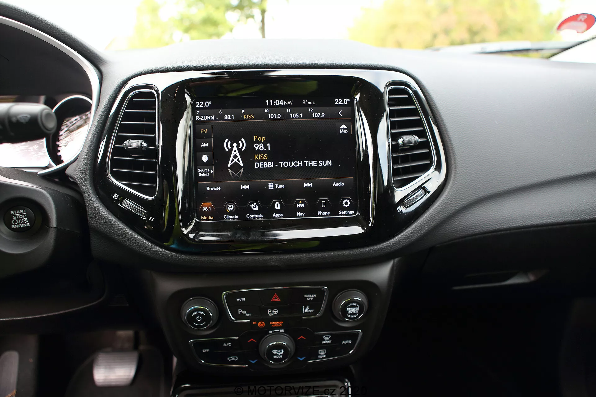 L'interno di una Jeep Compass del 2017, con particolare attenzione all'area centrale del cruscotto con un ampio touchscreen per l'infotainment montato centralmente che mostra funzionalità di connettività come radio, media e navigazione satellitare. Sotto il touchscreen ci sono i controlli del sistema di climatizzazione, inclusi quadranti e pulsanti con accenti cromati e un display LCD. I comandi montati sul volante parzialmente visibile includono opzioni per il controllo della velocità di crociera e la regolazione del volume audio. Sono mostrati anche dettagli di design aggiuntivi come le prese d'aria verticali, i comandi di riscaldamento dei sedili e una porta USB, mettendo in evidenza le caratteristiche tecnologiche interne e il design estetico del veicolo.