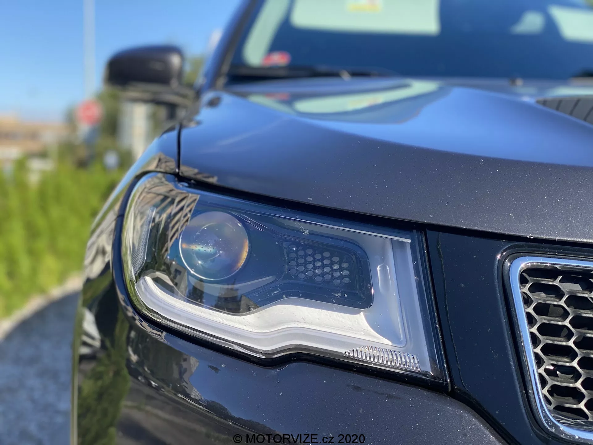 Primo piano dell'angolo anteriore sinistro di un SUV contemporaneo, con focus sul gruppo ottico avanzato con fascio principale a proiettore e luci di marcia diurna a LED. L'immagine mostra parte del design della griglia a nido d'ape, carrozzeria nera o scura con design robusto e contornato, e una vista parziale di un grande specchietto laterale sotto luce diurna con buone condizioni di illuminazione. Vegetazione sfocata in primo piano aggiunge profondità e un tocco artistico.