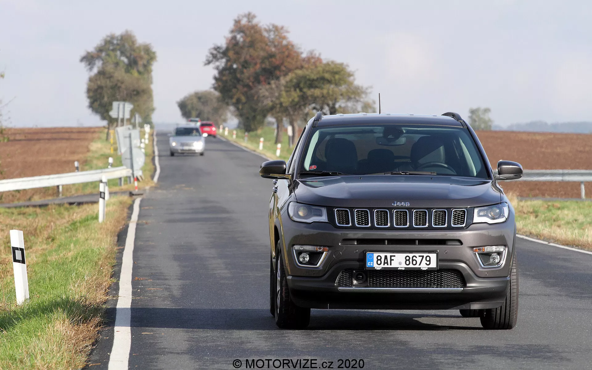 Inquadratura a bassa angolazione di una Jeep Compass 2017 che guida verso la fotocamera su una strada asfaltata a due corsie. La vista frontale mostra la griglia, i fari e il paraurti in dettaglio con caratteristiche moderne visibili come luci LED o HID e sensori di parcheggio anteriori. L'auto è in movimento, con uno sfondo rurale sfocato che presenta pali bianchi lungo la strada e campi aperti sotto un cielo sereno.