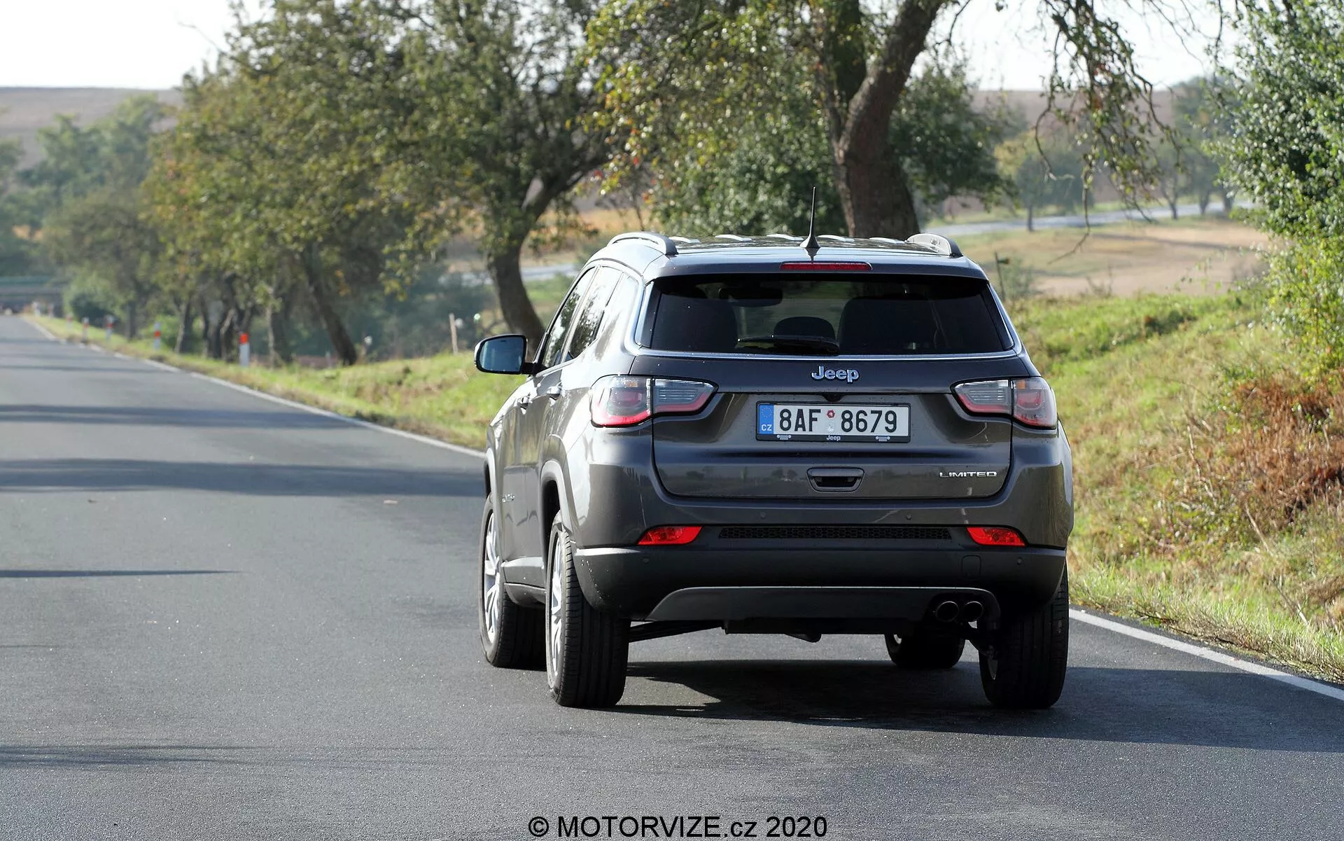 Vista posteriore di una Jeep Compass SUV del 2017 grigio metallizzato che si allontana su una strada a due corsie, posizionata leggermente a destra del fotogramma. Il veicolo, nella corsia di destra, presenta caratteristiche visibili come il portellone con il distintivo Jeep e la targa, il lunotto posteriore con un piccolo spoiler, luci posteriori con sezioni rosse e bianche, paraurti posteriore con sensori e un'antenna a pinna di squalo sul tetto. Le ruote in lega sono parzialmente visibili. La scena suggerisce una giornata soleggiata con alberi e erba circostanti, ad indicare un'ambientazione rurale o semi-rurale.