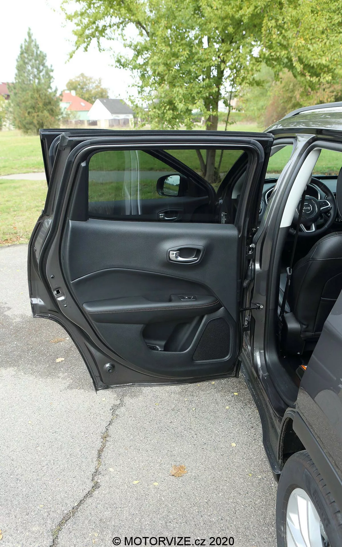 Vista interna di una Jeep Compass 2017 dall'esterno che mostra la porta lato conducente aperta con una maniglia metallica ed ergonomica, scomparti portaoggetti, griglia dell'altoparlante e pannello di controllo per le operazioni di finestra e serratura della porta. Sono visibili parte del cruscotto, della console centrale e del volante multifunzione, suggerendo un interno moderno e ricco di funzionalità. Luce naturale, clima coperto, alberi sullo sfondo suggeriscono un ambiente all'aperto in un parco o su una strada alberata.