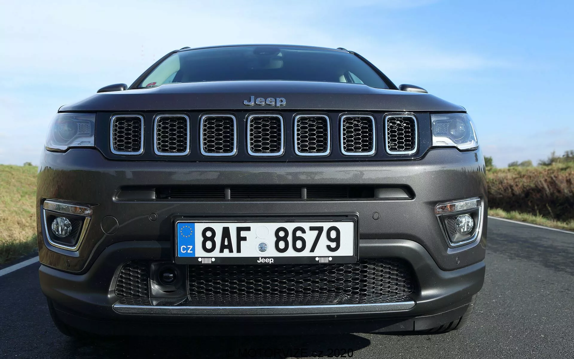 Vista frontale dal basso di una Jeep Compass 2017, che enfatizza la caratteristica griglia a sette feritoie e il paraurti anteriore centrale con una grande griglia di aspirazione dell'aria. La foto ravvicinata, leggermente fuori centro a destra, mette in risalto la posizione imponente del veicolo con un focus sulla griglia, sui dettagli del paraurti, sulla targa in stile europeo e sugli elementi di design robusti, evidenziando l'identità e la capacità fuoristrada della Jeep.