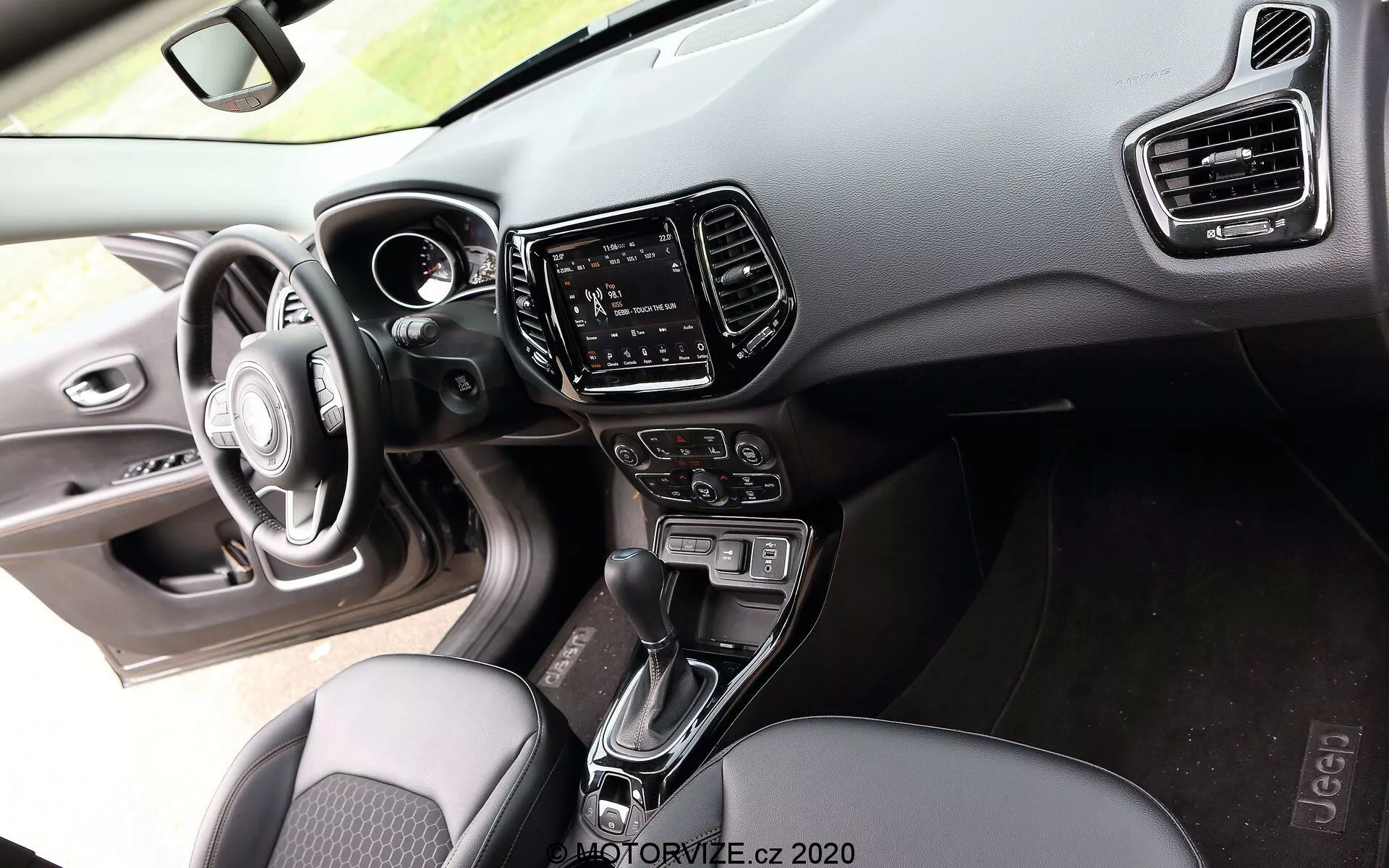 Vista interna di una Jeep Compass 2017 dalla prospettiva del passeggero, che mostra un volante a tre razze con pulsanti di controllo integrati, un cruscotto digitale parzialmente visibile sul cruscotto realizzato con materiali soft-touch e plastica dura con cuciture visibili, un sistema di infotainment touch-screen nella console centrale fiancheggiato da bocchette di climatizzazione verticali, una leva di cambio automatico tra i sedili anteriori, pulsanti e manopole per il controllo del clima, sedili in tessuto con un design a motivo e uno scorcio del pannello porta del conducente con controlli di regolazione dei finestrini e degli specchietti laterali esaltati dall'illuminazione ambientale, mettendo in evidenza l'attenzione al comfort e alla tecnologia nel mercato di fascia media a alta.