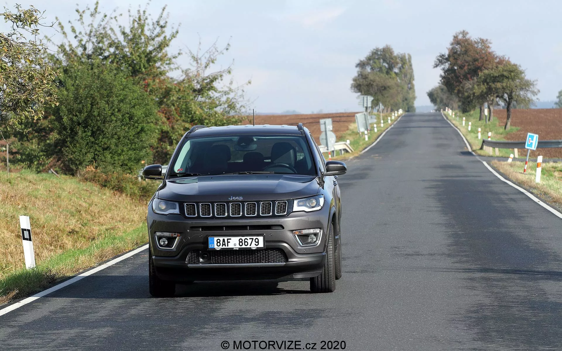 Vista frontale di una Jeep Compass 2017 che viaggia a velocità moderata su una strada di campagna. L'immagine cattura le caratteristiche frontali distintive tra cui la iconica griglia frontale a sette slot, fari rettangolari moderni e fendinebbia integrati nel paraurti. L'auto è fotografata a livello degli occhi, evidenziando il design esterno e la tecnologia di illuminazione in buone condizioni di luce naturale. Una targa in stile europeo è montata centralmente nella parte anteriore, con un chiaro scenario rurale sullo sfondo.
