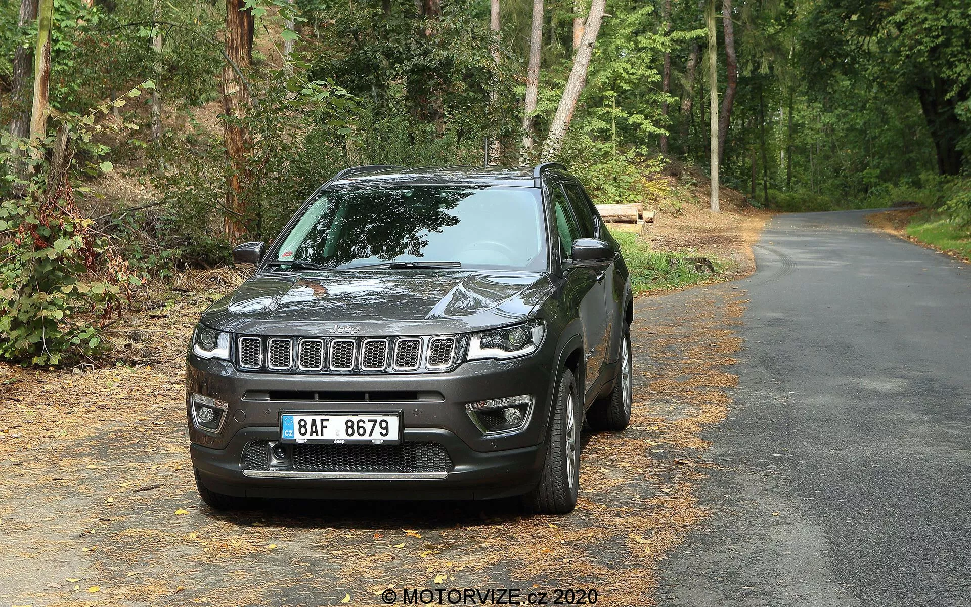 SUV Jeep Compass 2017 su strada asfaltata circondata da vegetazione, posizionato di fronte alla fotocamera con un angolo leggermente a sinistra mostrando la parte anteriore e il lato del conducente. Le caratteristiche visibili includono una griglia frontale a listelli orizzontali con emblema centrale, fari rettangolari a LED, alloggiamenti per fari fendinebbia nel paraurti inferiore, cofano sagomato che riflette luce, calotte degli specchietti in tinta con la carrozzeria, finestre leggermente oscurate, cerchio in lega a più razze e parafango delineato. Sullo sfondo alberi e fogliame indicano un'area stradale alberata.